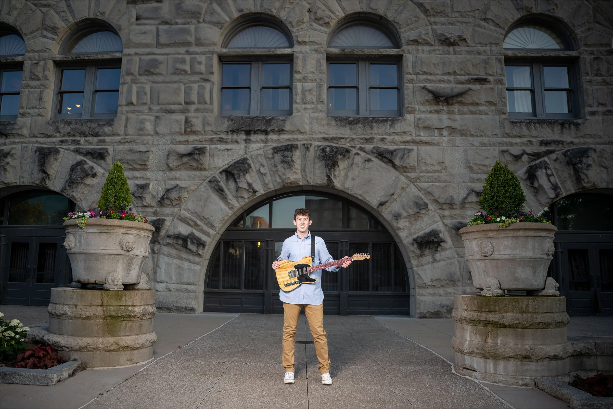 A young man dressed in a blue shirt and khaki pants is standing in front of a stone building with large planters of flowers on either side. He is holding an electric guitar and smiling.