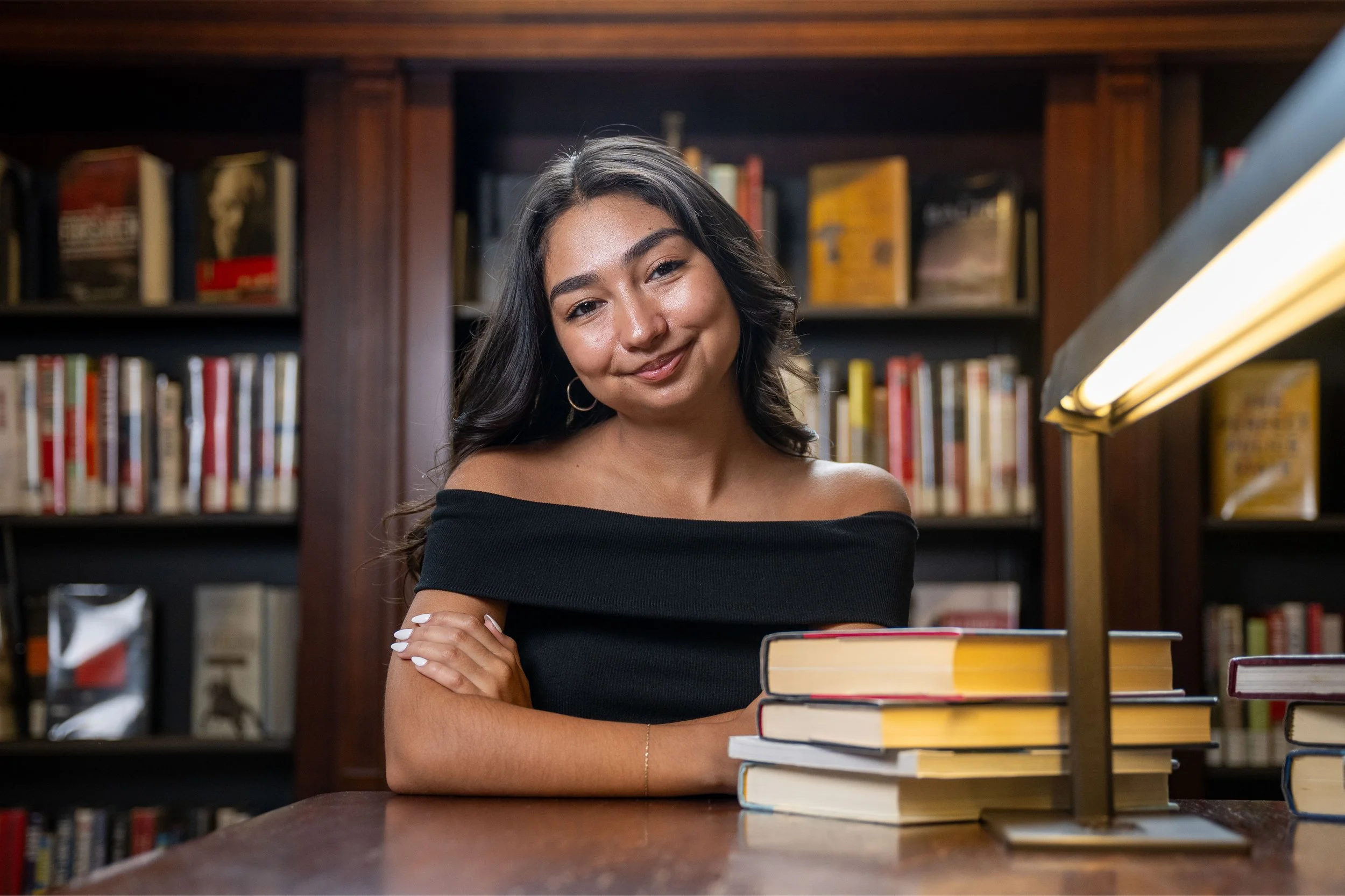 A woman with dark wavy hair and hoop earrings smiling at the camera in a library, sitting at a table with a stack of books in front of her, surrounded by bookshelves filled with various books.