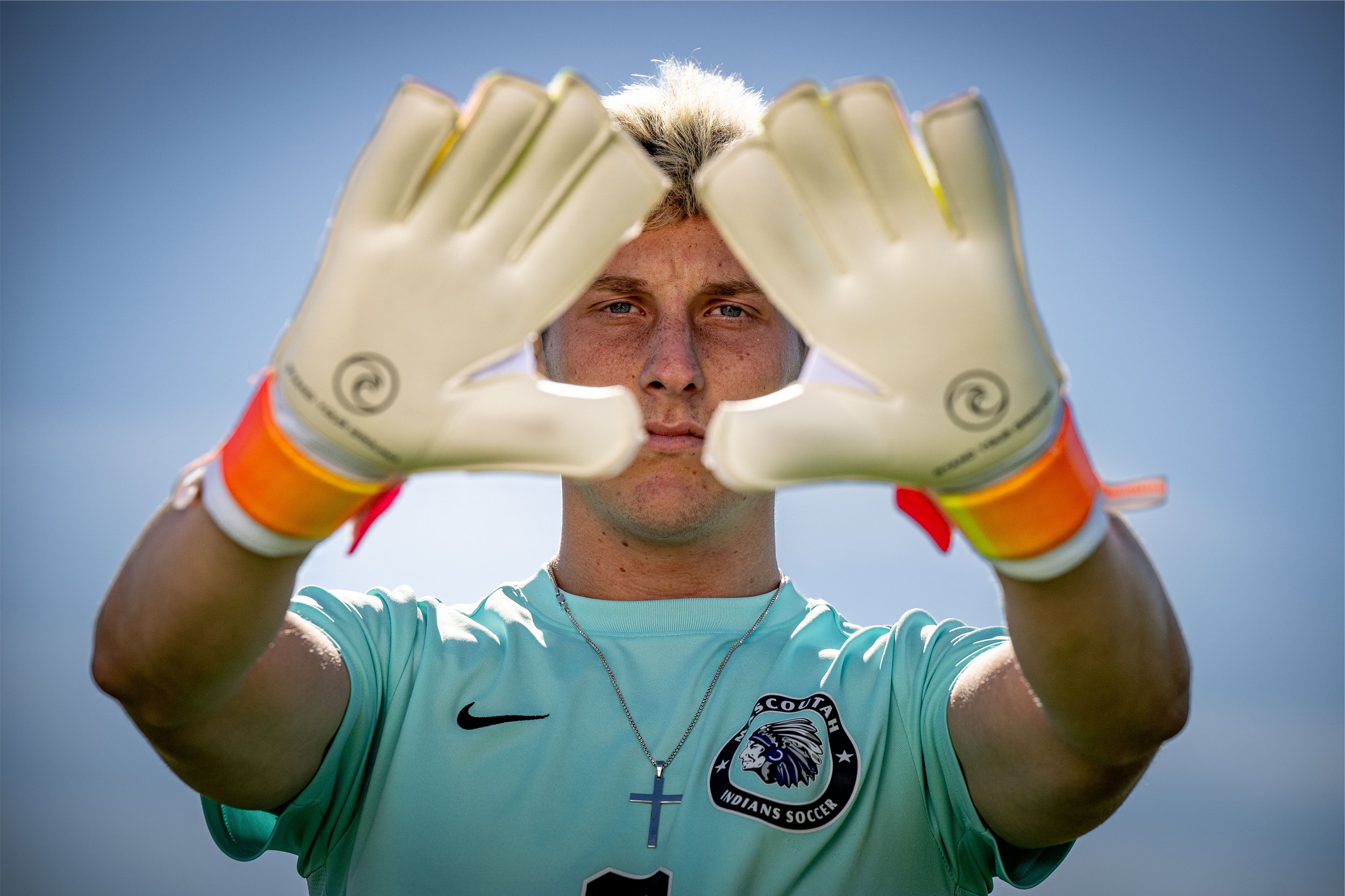A young male soccer goalkeeper with blond hair wearing a light blue jersey with a team logo, holding his hands in front of his face with white goalkeeper gloves, and looking through the gap between his gloves against a blue sky background.