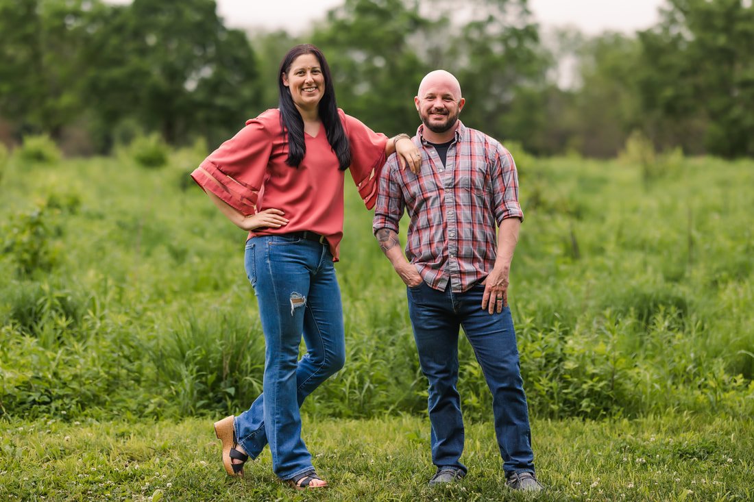 A woman and a man standing outdoors on grass, smiling, with green trees in the background.