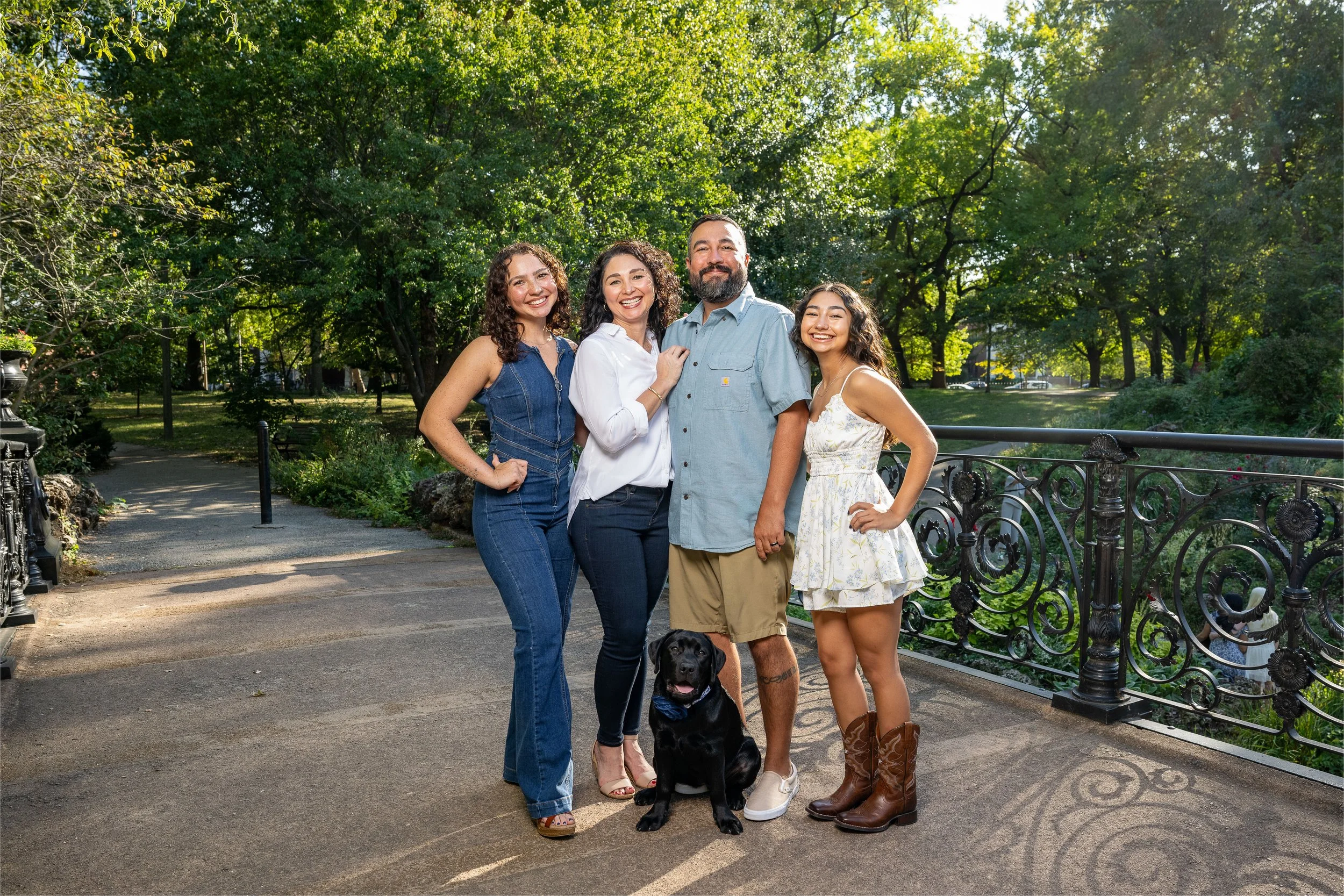 A group of five people and a dog posing for a photo on a pathway in a park with trees in the background. The group includes four women and one man, all smiling and dressed in casual summer clothing.