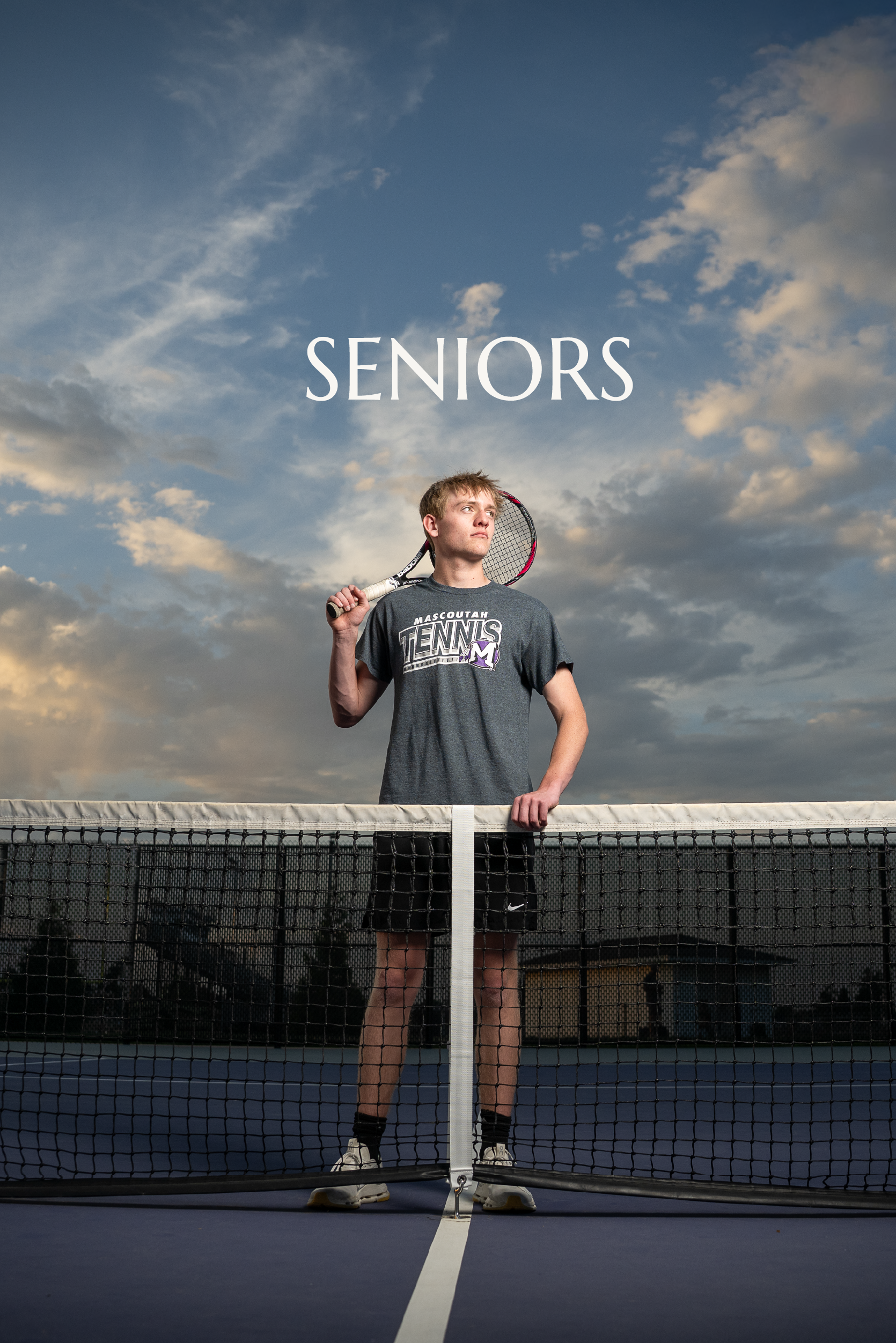 A young man standing on a tennis court, holding a tennis racket over his right shoulder, wearing a 'Mascoutah Tennis' t-shirt, behind a tennis net, with a cloudy sky backdrop.