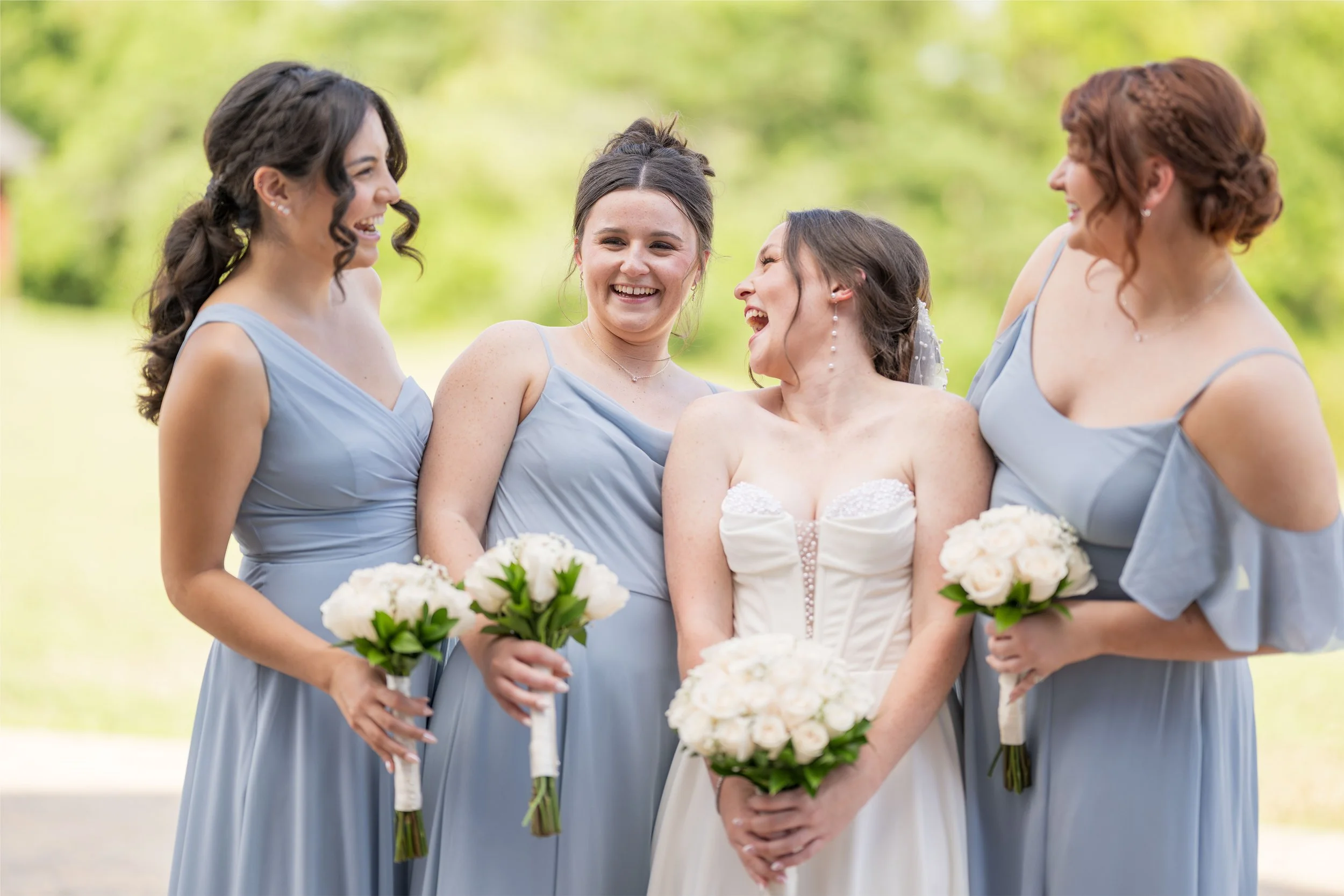 A group of five women dressed in light blue dresses and a white wedding gown, holding bouquets of white roses, standing outdoors with a lush green background, smiling and engaging in conversation.