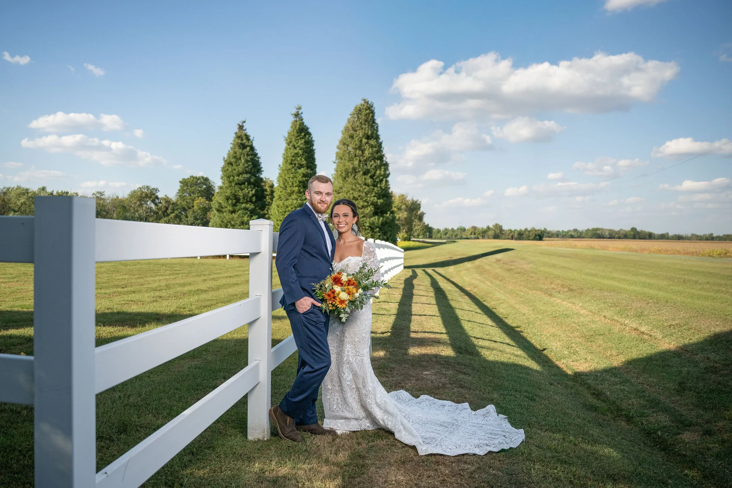 A newlywed couple standing outdoors alongside a white fence under a blue sky with scattered clouds. The groom is wearing a blue suit, and the bride is in a white lace wedding dress holding a bouquet of sunflowers and other flowers. They are smiling a