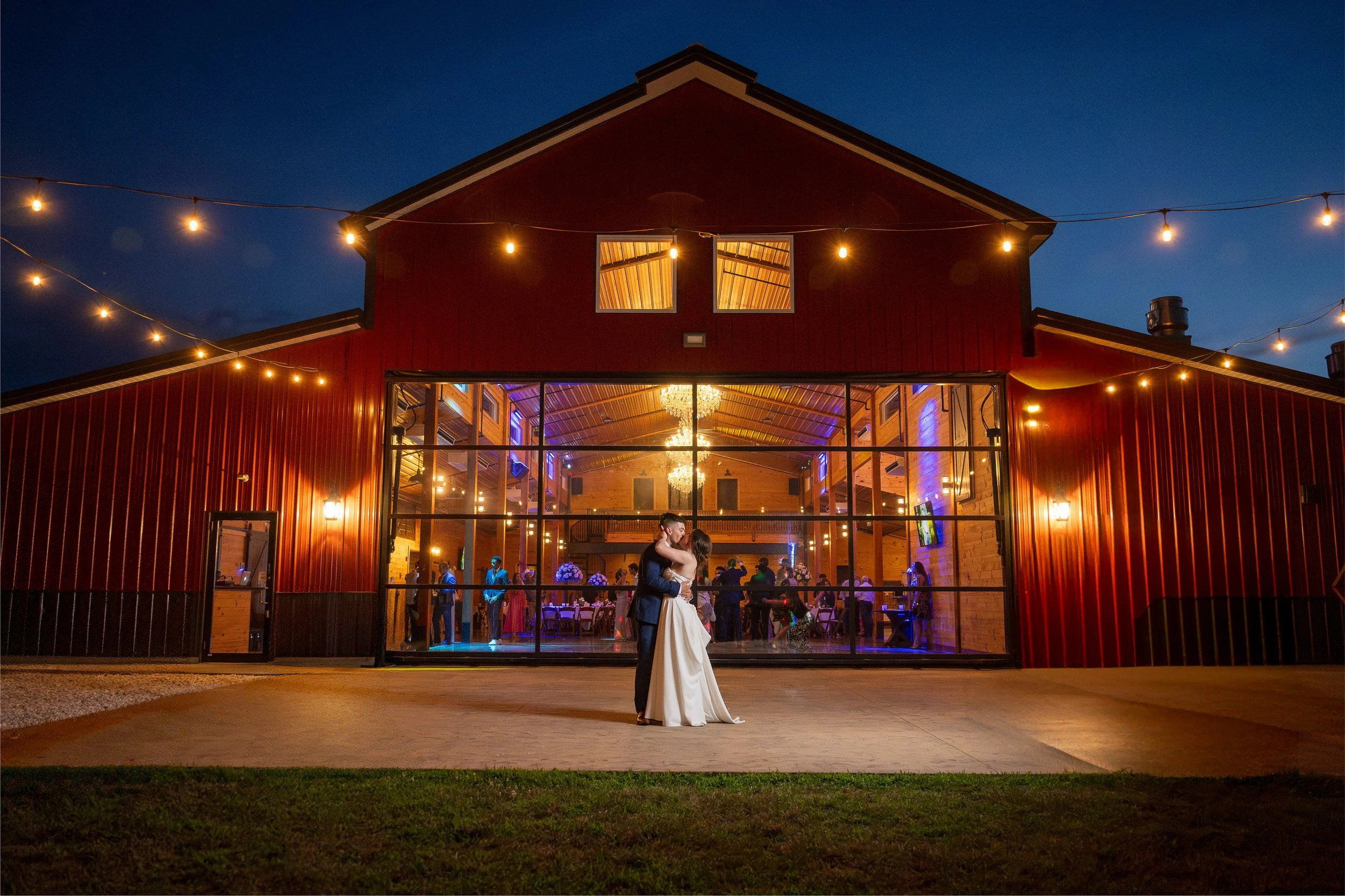 A bride and groom dancing outside a rustic barn decorated with string lights during their wedding reception at night.
