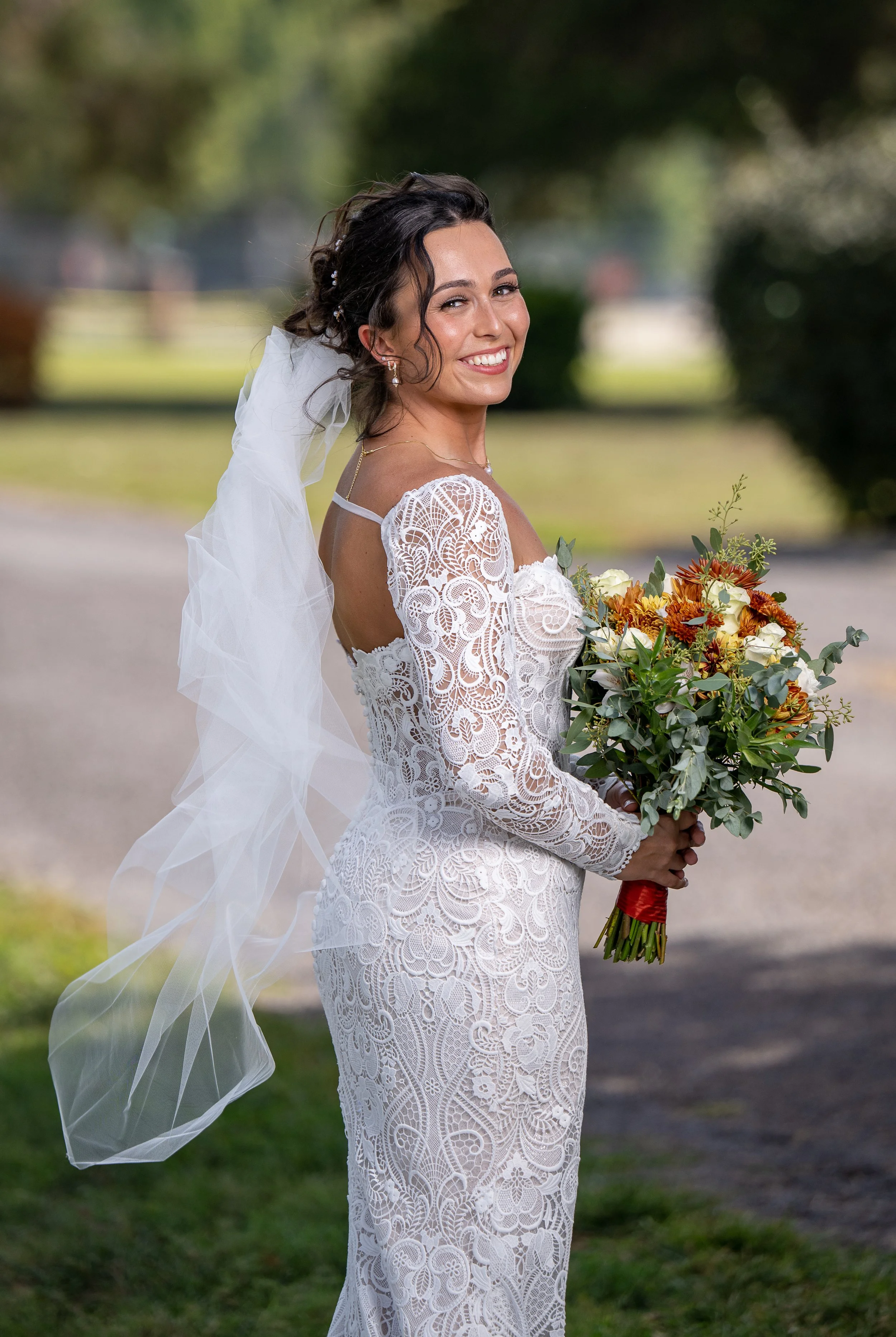 A smiling bride with dark hair styled with small pearl accessories, wearing a white lace wedding dress with long lace sleeves, holding a bouquet of white, red, and orange flowers with green foliage, outdoors on a sunny day with blurred trees in the b
