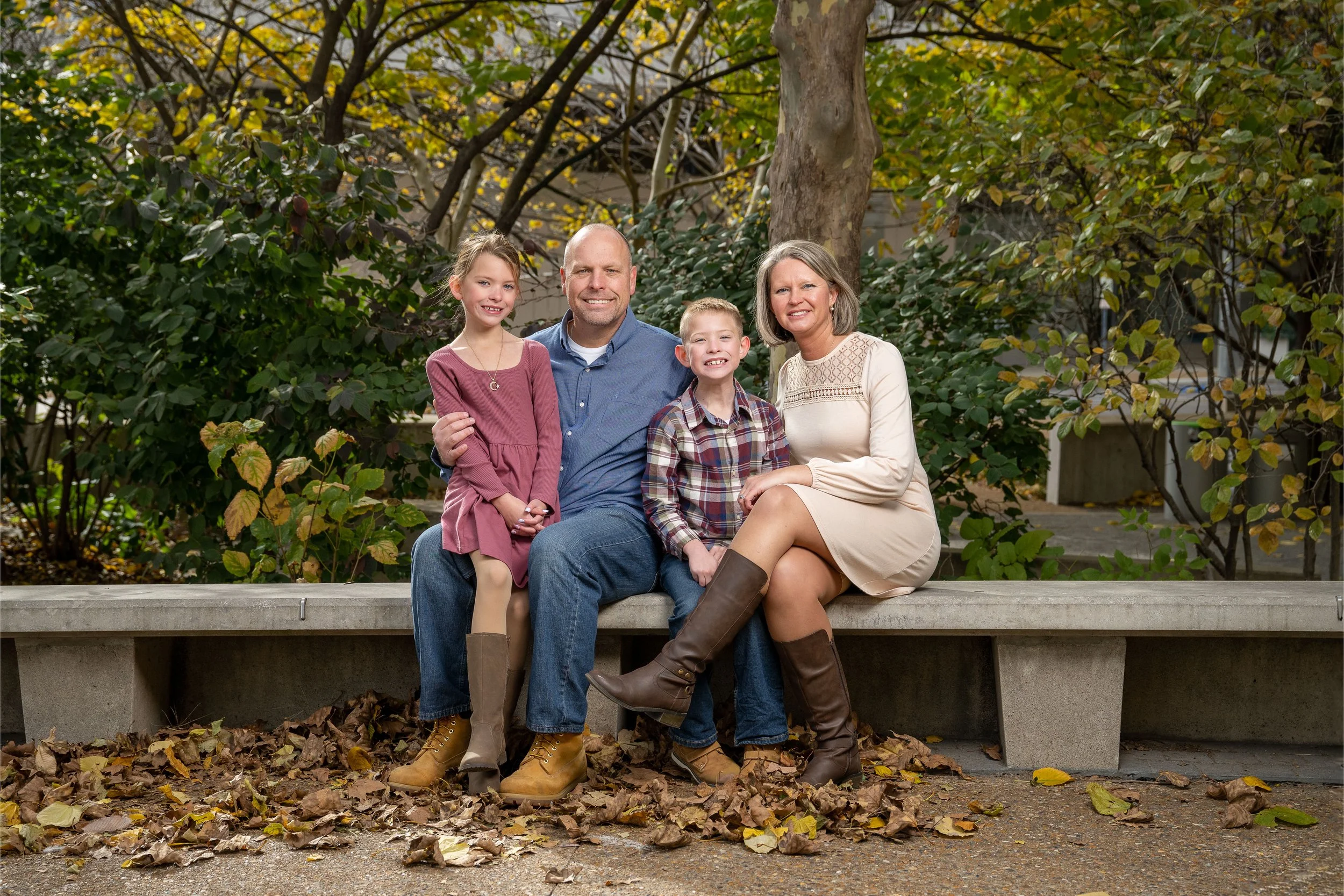 A family of four sitting on a concrete bench in a park during autumn, surrounded by fallen leaves and trees with green and yellow leaves. They are smiling and looking at the camera.