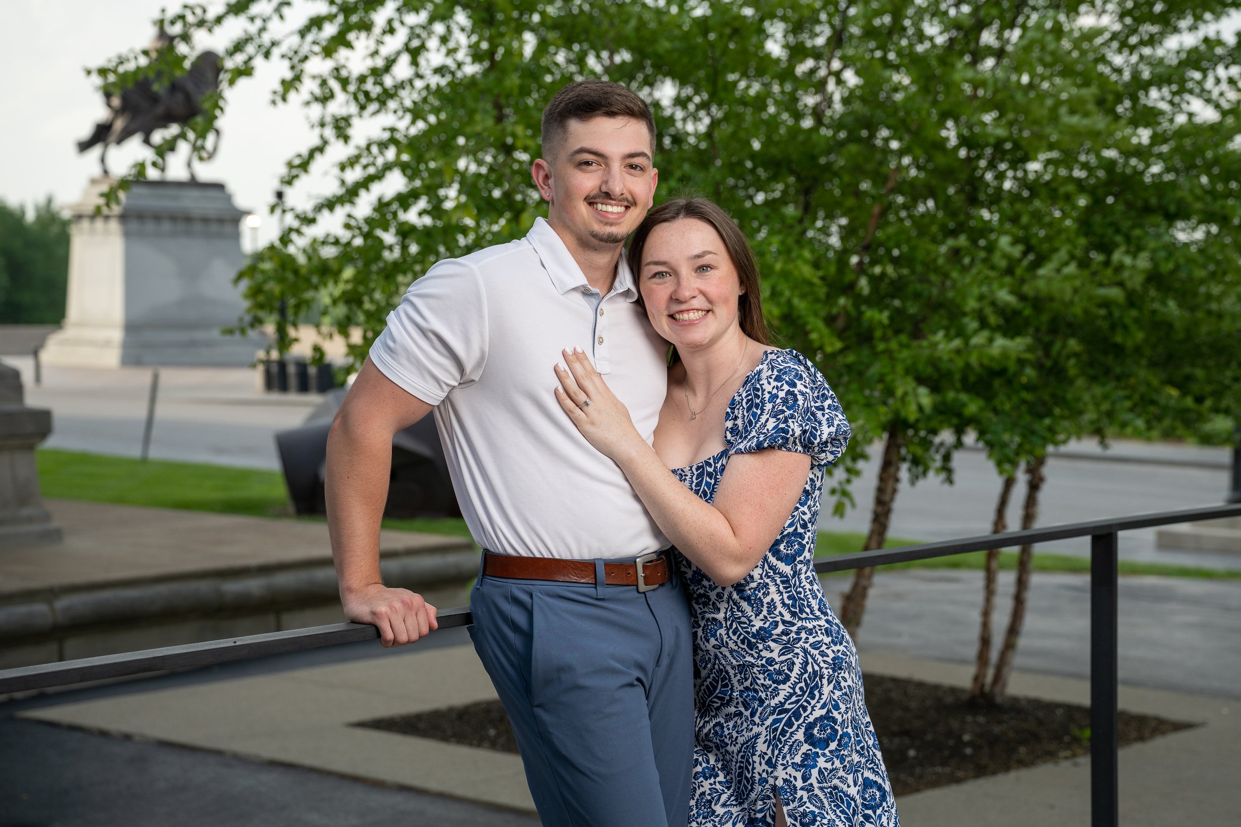 A young couple smiling and posing closely outdoors, with trees and a monument in the background.