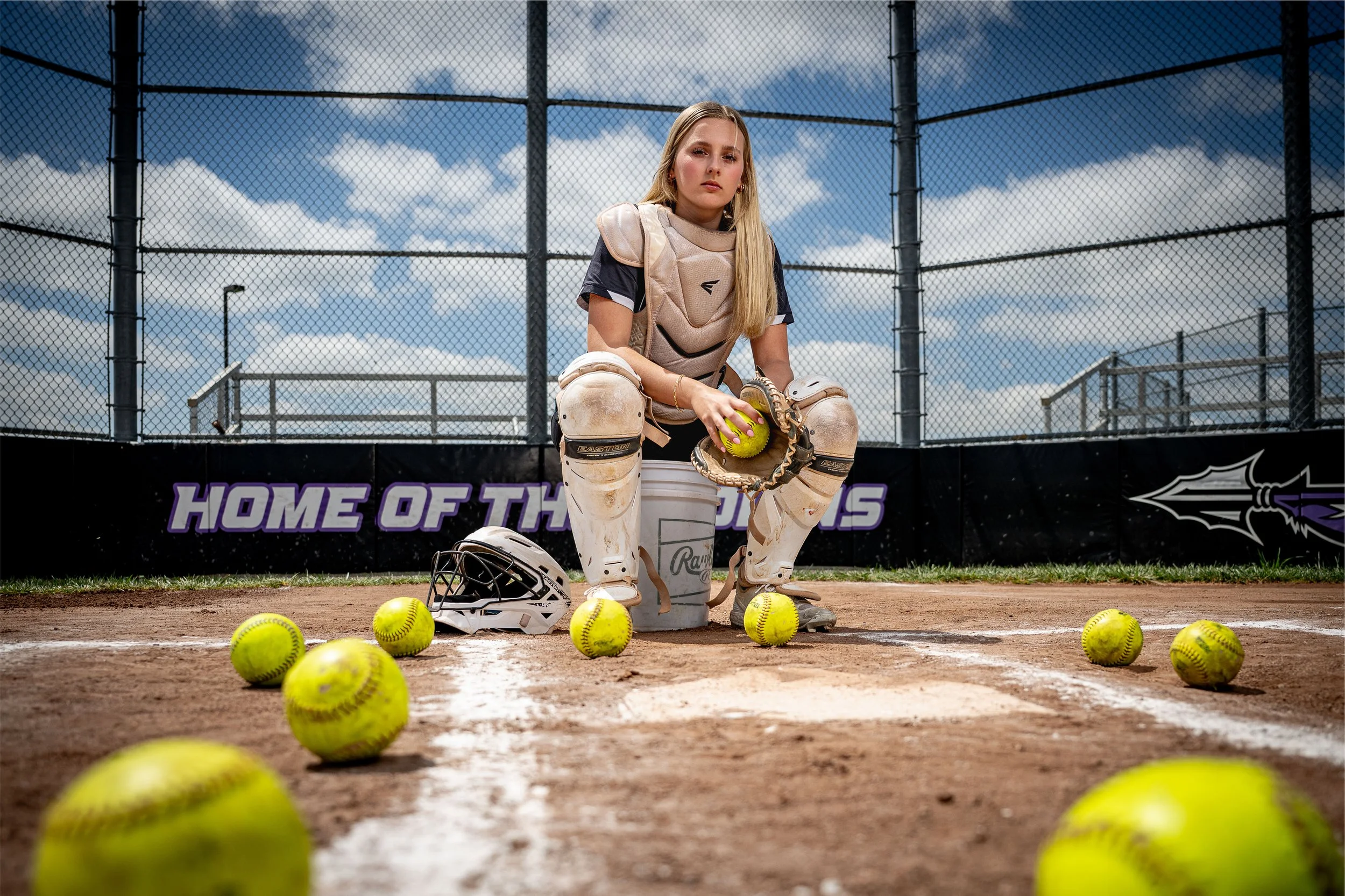 A young female softball goalie in protective gear, sitting on a dirt field with a cloudy sky background, surrounded by yellow softball balls, a helmet on the ground, and a bucket for the balls.