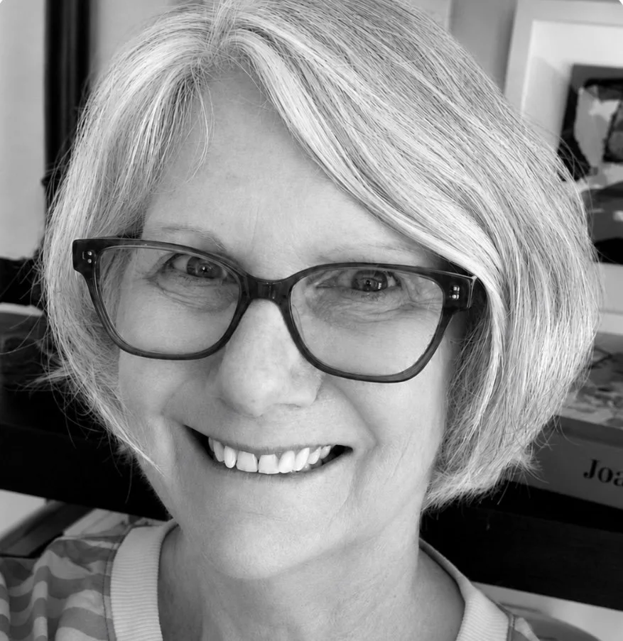 Close-up black and white portrait of a smiling woman wearing glasses and a patterned shirt, with a background of bookshelves and boxes.