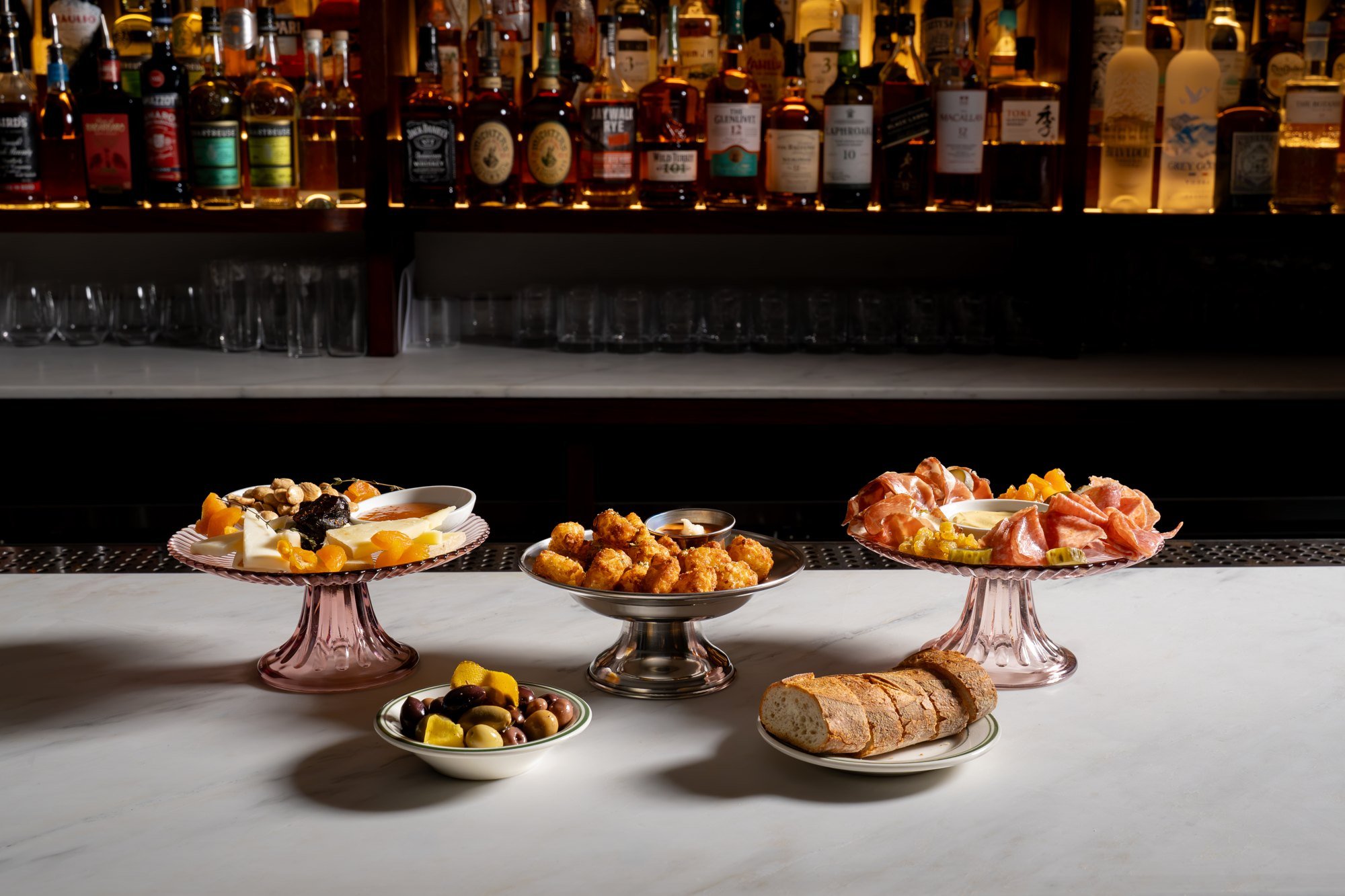 Bar top spread of snacks on pink glass cake stands in front of the back bar