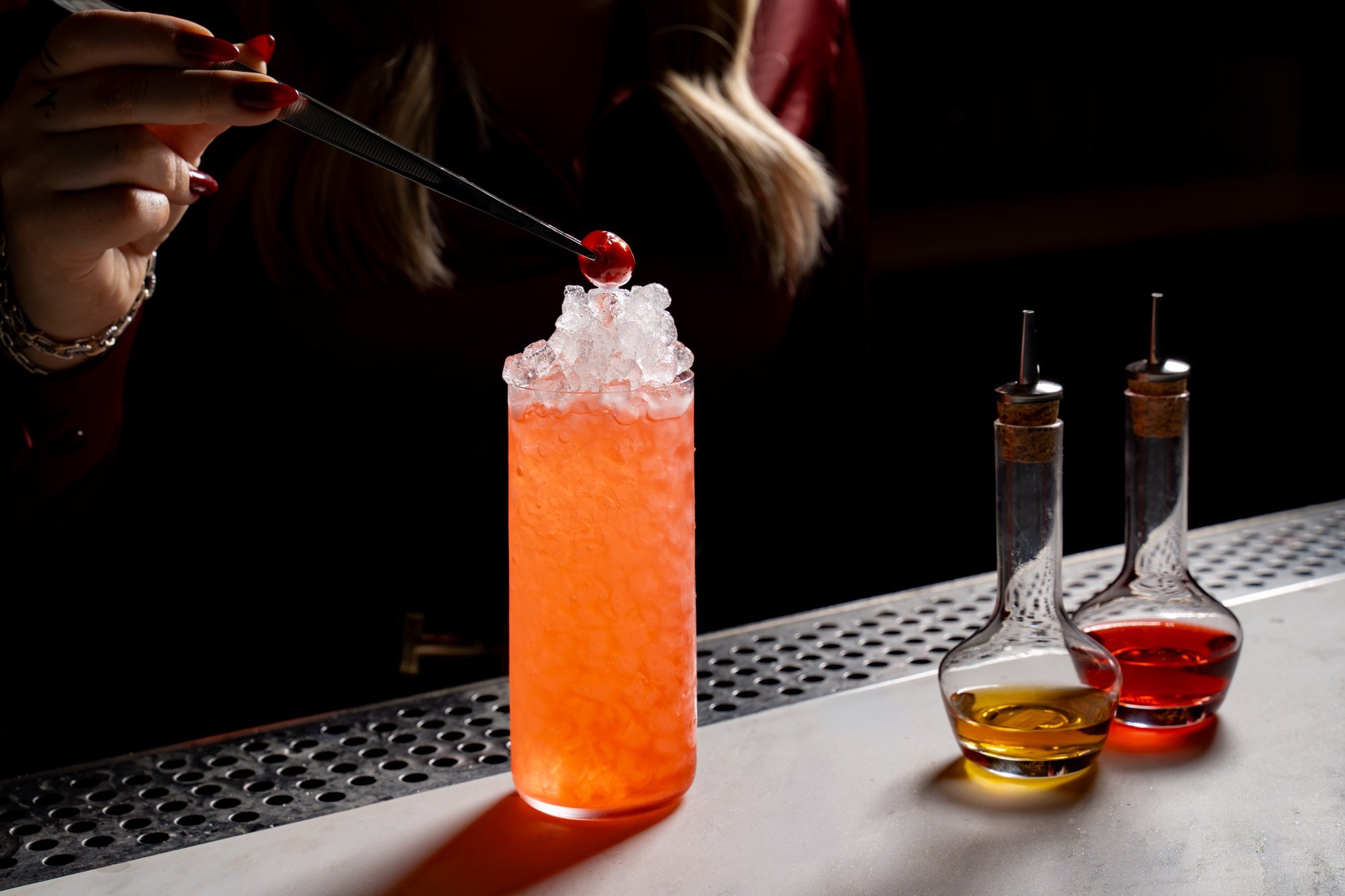 Bartender placing a cherry garnish on a red-orange cocktail over crushed ice