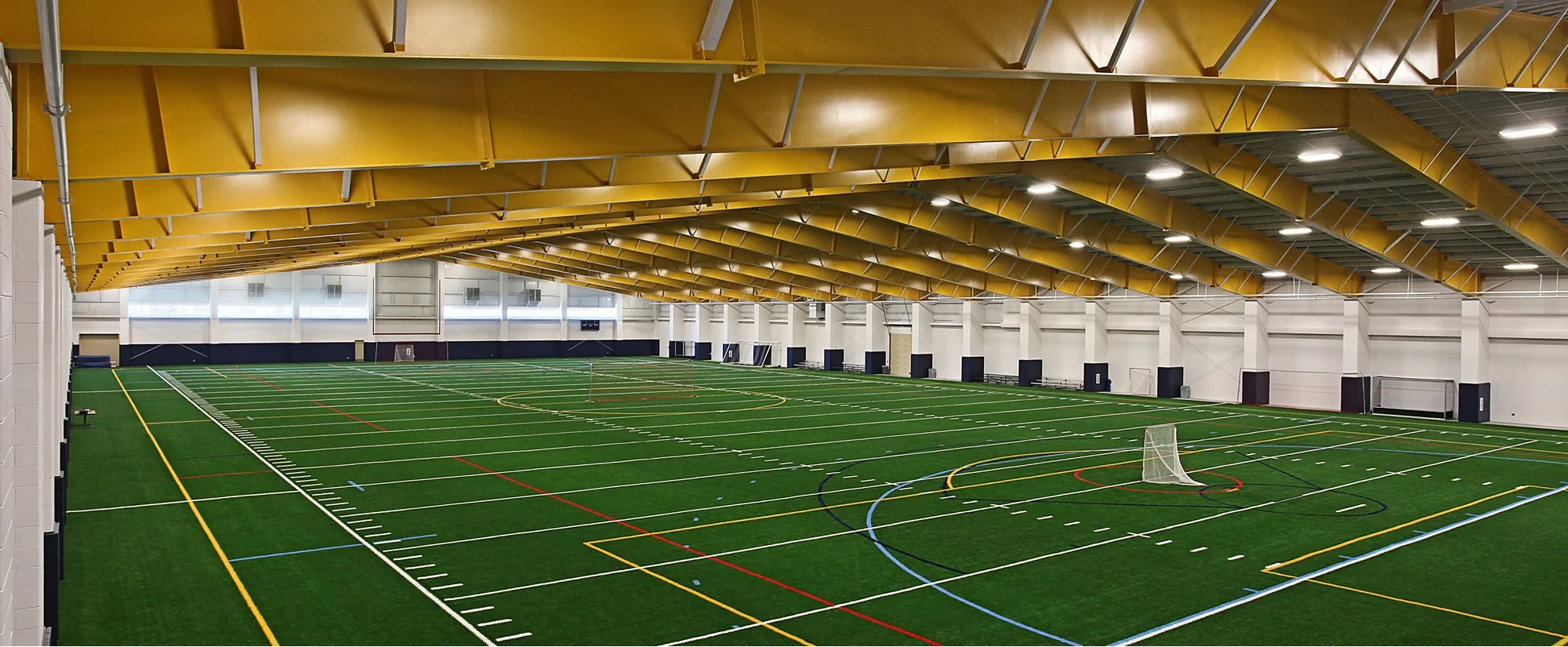 Indoor sports facility featuring a large artificial turf field with multiple colored lines for various sports. The ceiling has a yellow structural design with embedded lighting, and there are windows along the upper walls letting in natural light.