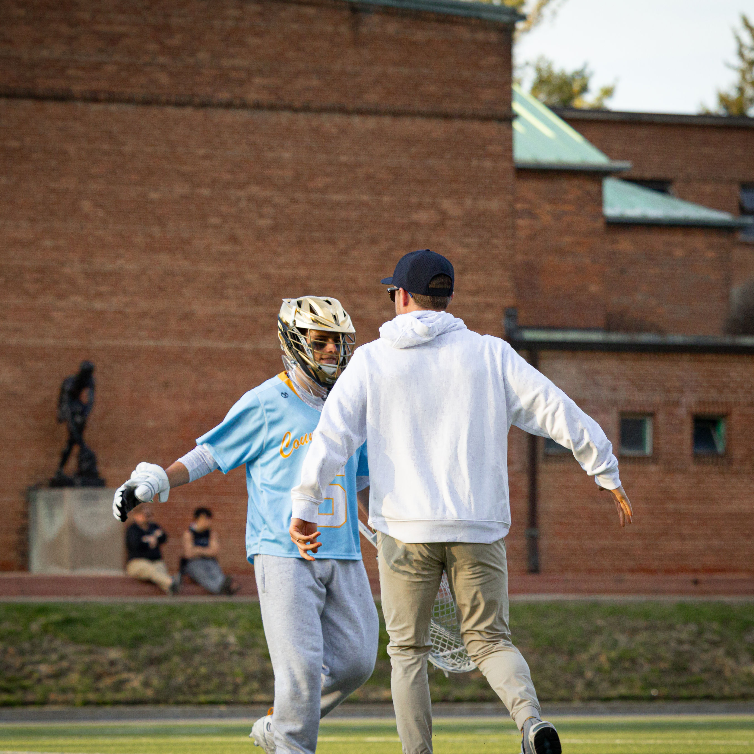 Two individuals wearing sports gear, including helmets and jerseys, are standing on a sports field facing each other, with a brick building and trees in the background.