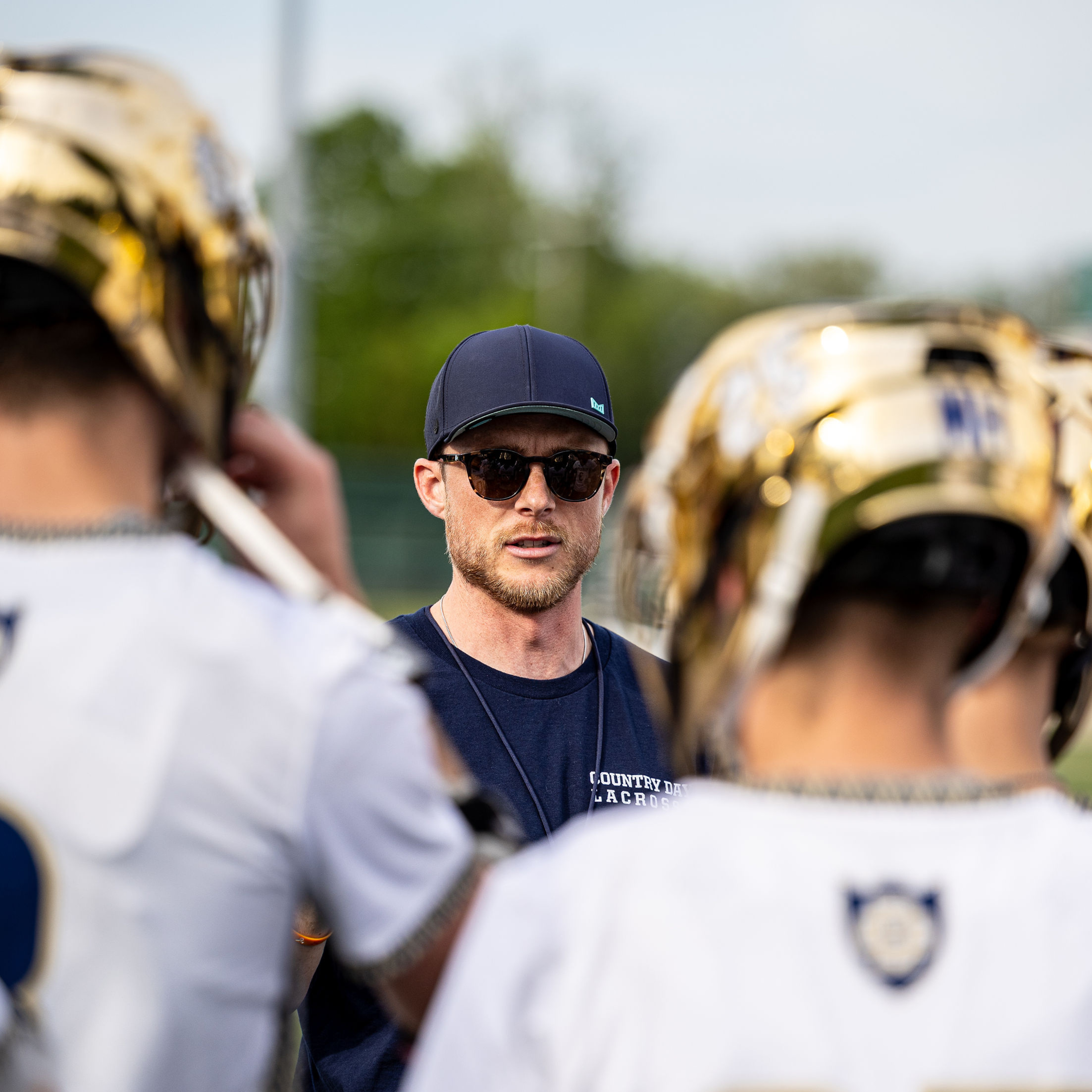 Close-up of a man with sunglasses and a dark blue cap standing outdoors, with multiple lacrosse players wearing gold helmets in the foreground and background.