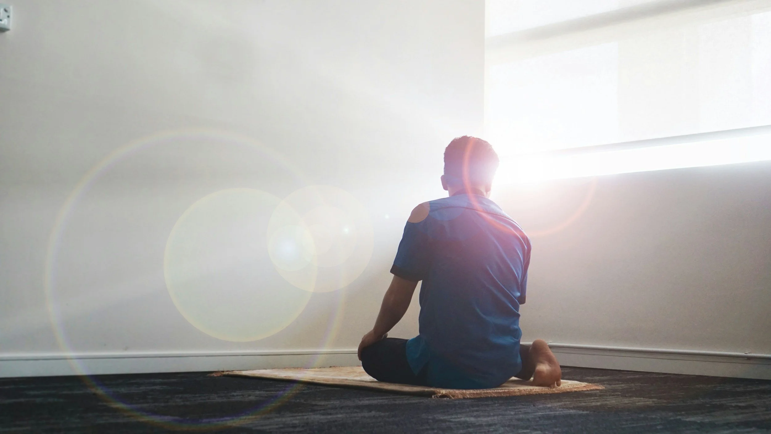 Person practicing yoga or meditation in a room with sunlight coming through a window, sitting on a yoga mat with legs crossed and back facing the window.