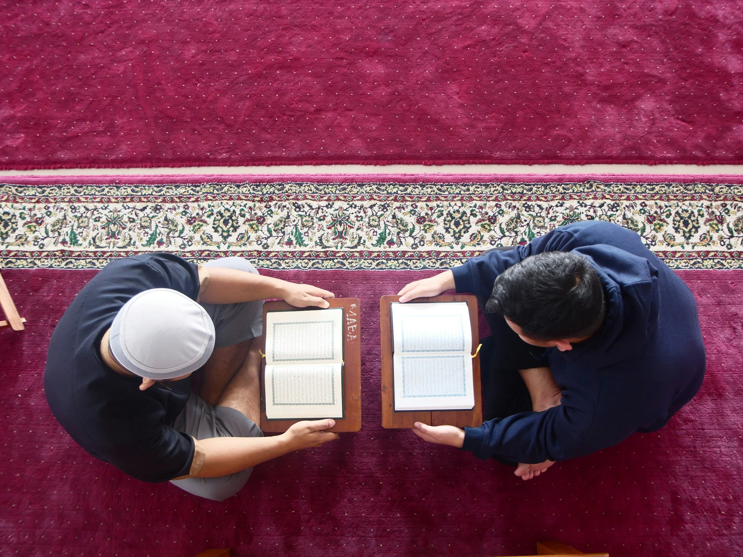 Two individuals sitting on a red carpet, facing each other, each holding open a religious book, with prayer mats beside them.