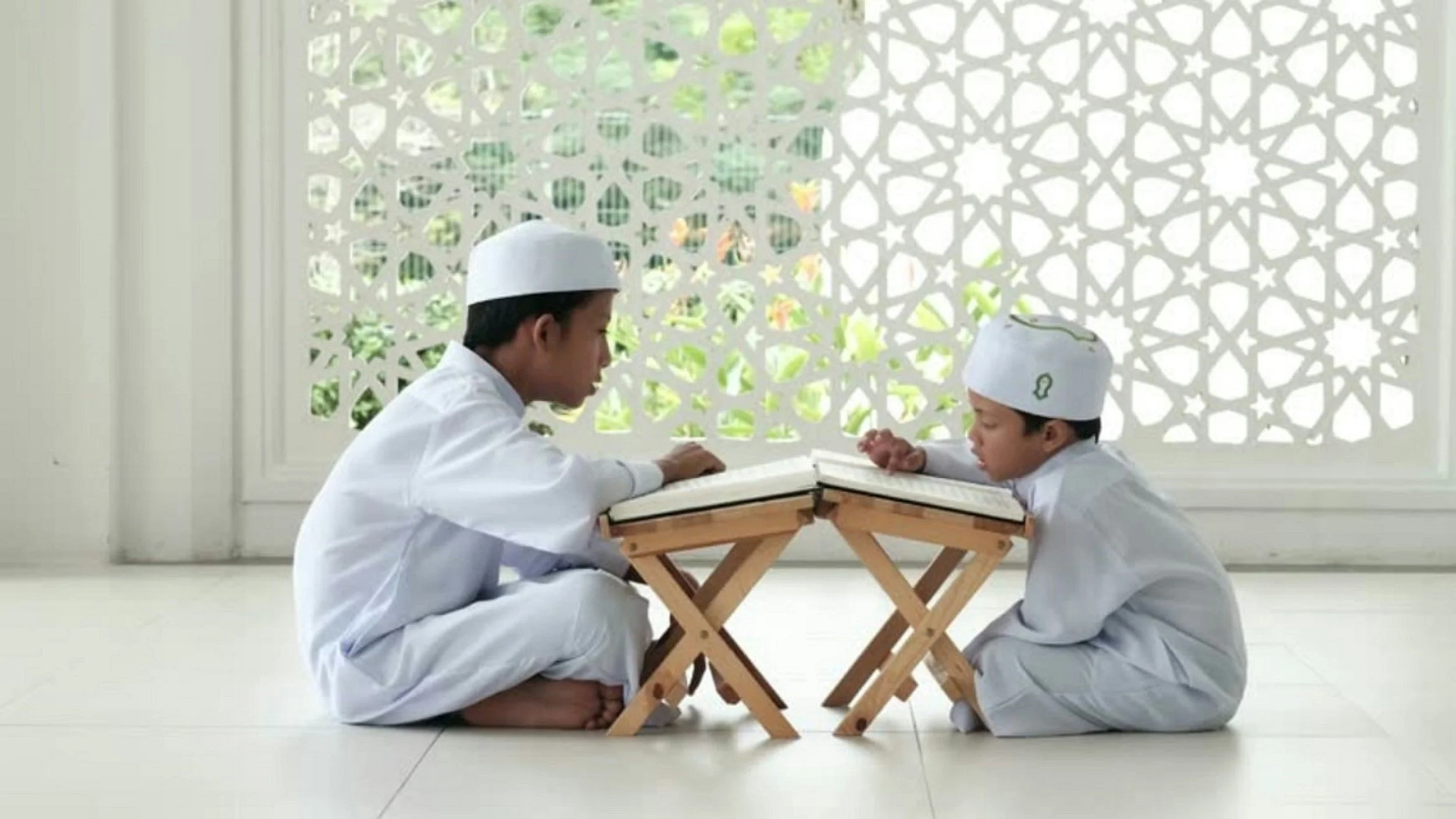 Two young boys, dressed in white traditional clothing and white caps, sit facing each other on the floor with a small wooden table between them, reading from a book. The background features an ornate white lattice wall with sunlight filtering through.
