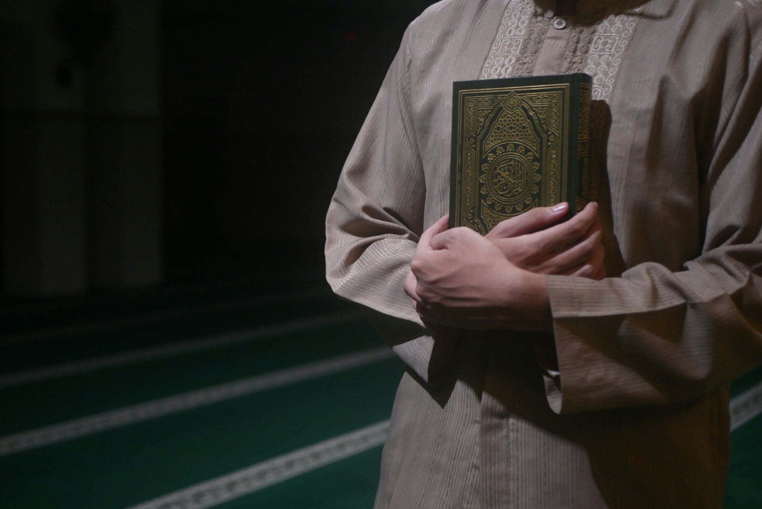 Person holding a green and gold patterned book in their arms, wearing a beige traditional garment, in a dimly lit space.