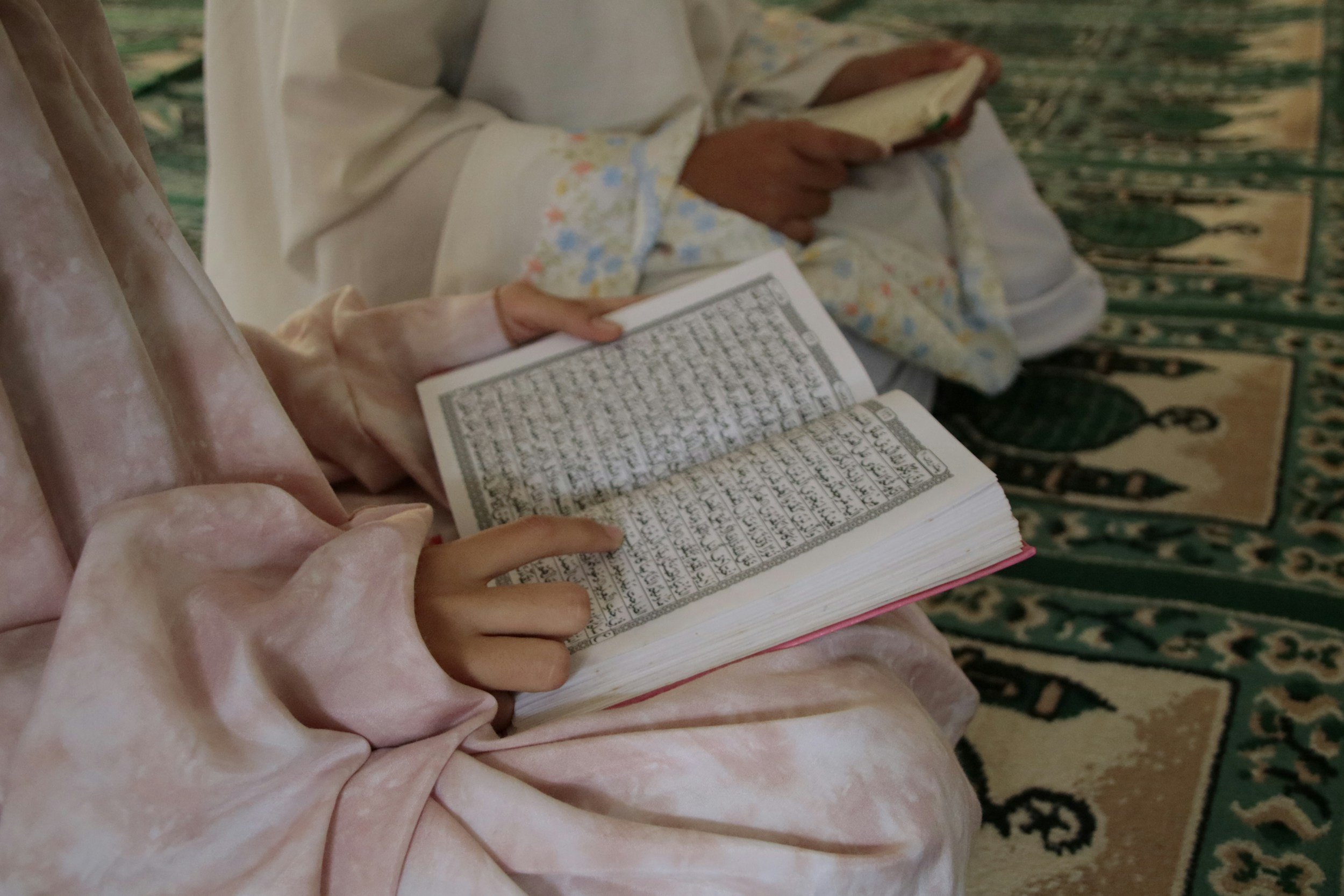 Close-up of a person reading the Quran, with someone else in the background holding their phone, sitting on a green patterned carpet.