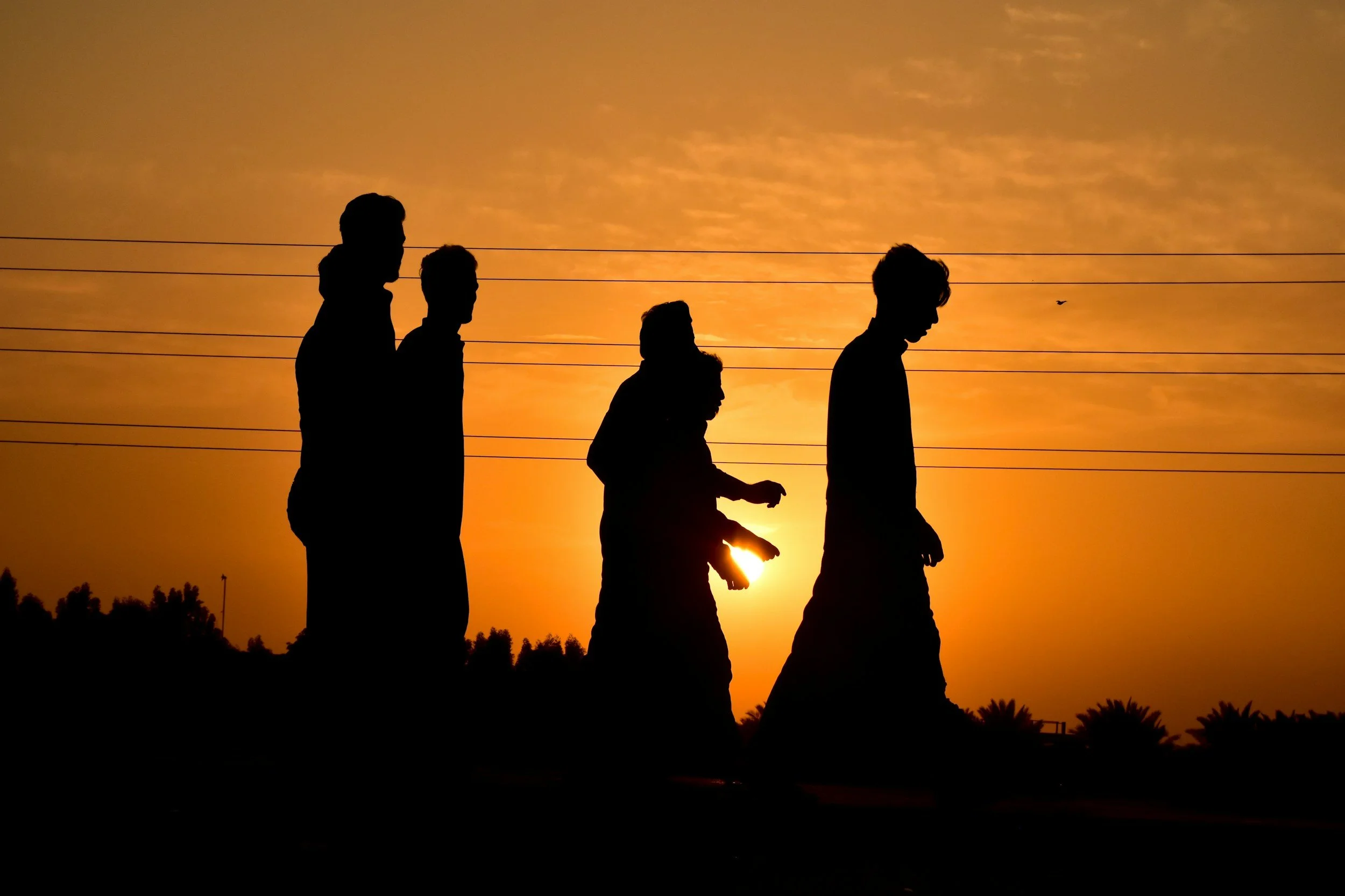 Silhouettes of four people walking outdoors during sunset with a bright orange sky, power lines, and trees in the background.