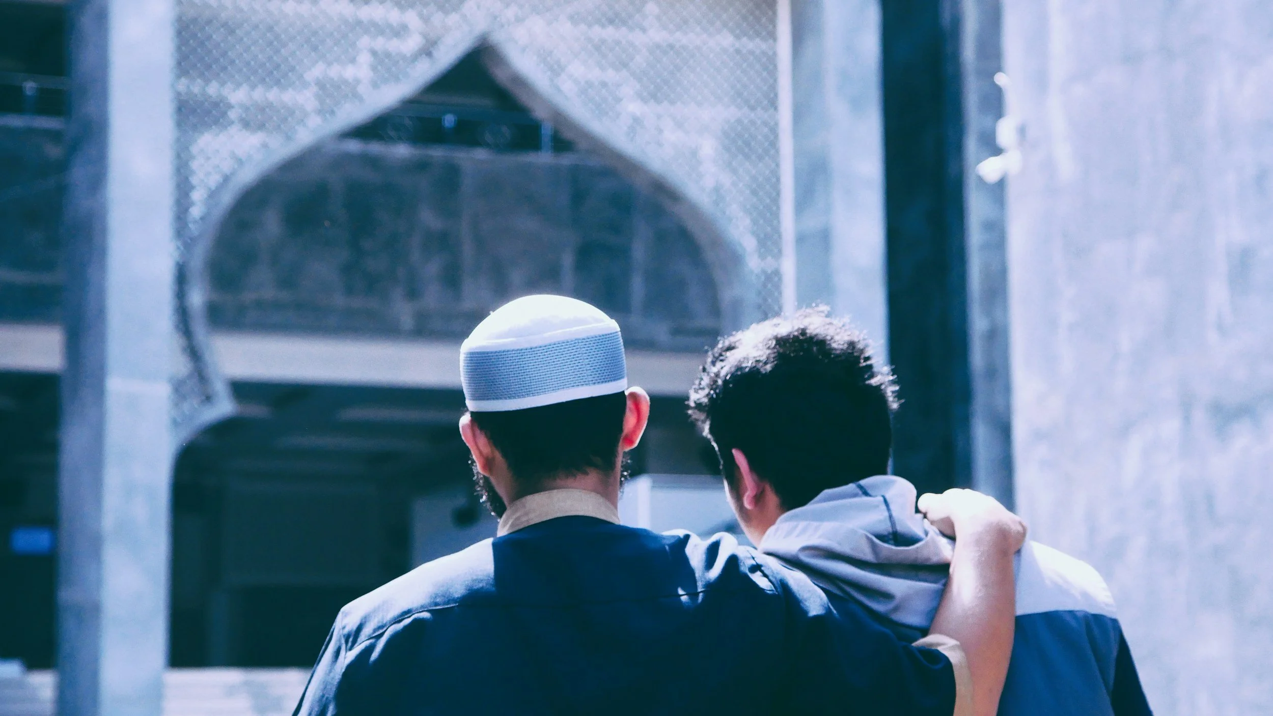 Two men are standing together, looking up at a mosque's minaret. One is wearing a white skullcap and the other has curly hair, and they are embracing each other.