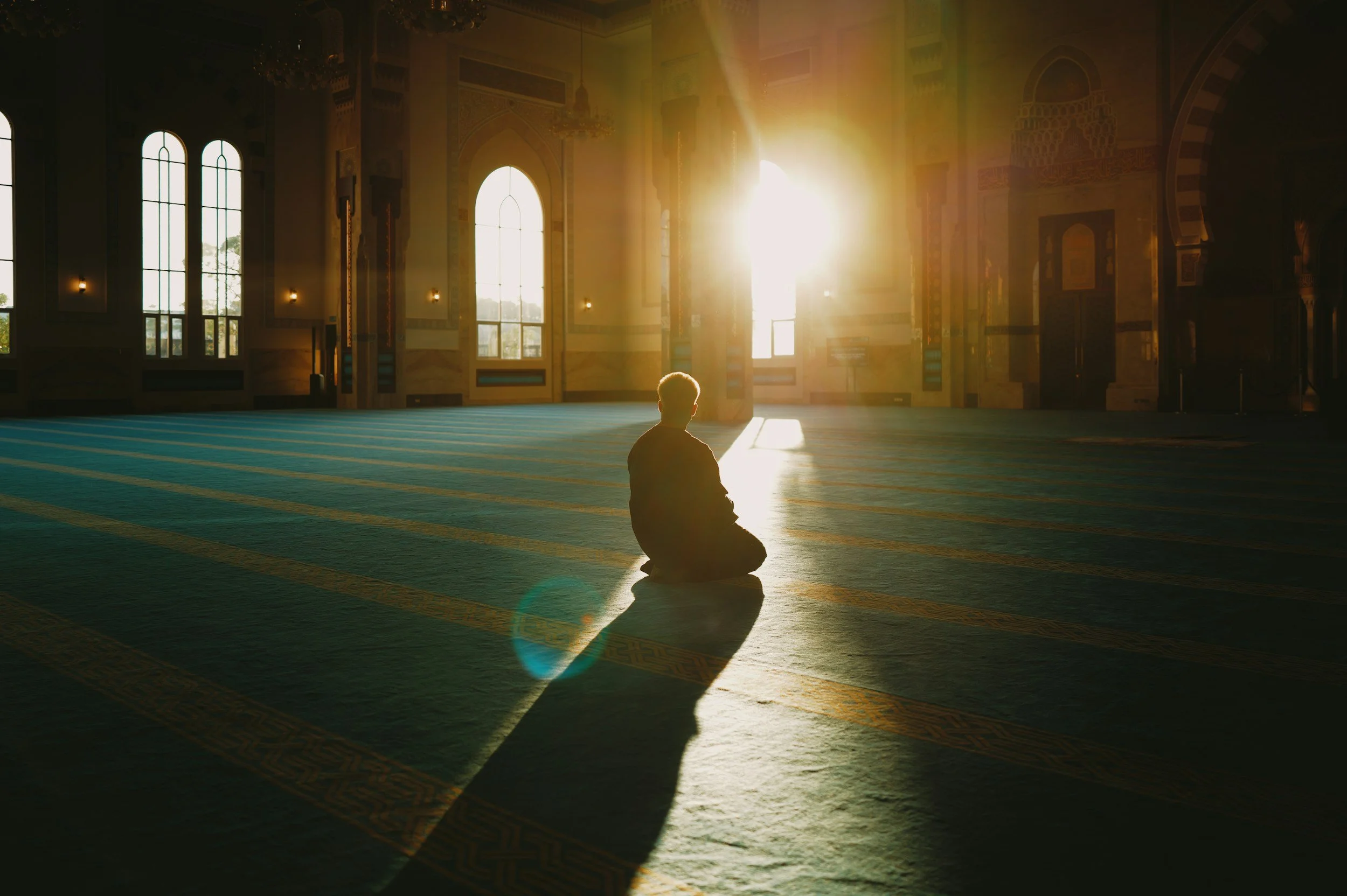 A person sitting in a prayer position inside a mosque during sunrise or sunset, with sunlight streaming through large windows, illuminating the prayer rug and creating a peaceful atmosphere.
