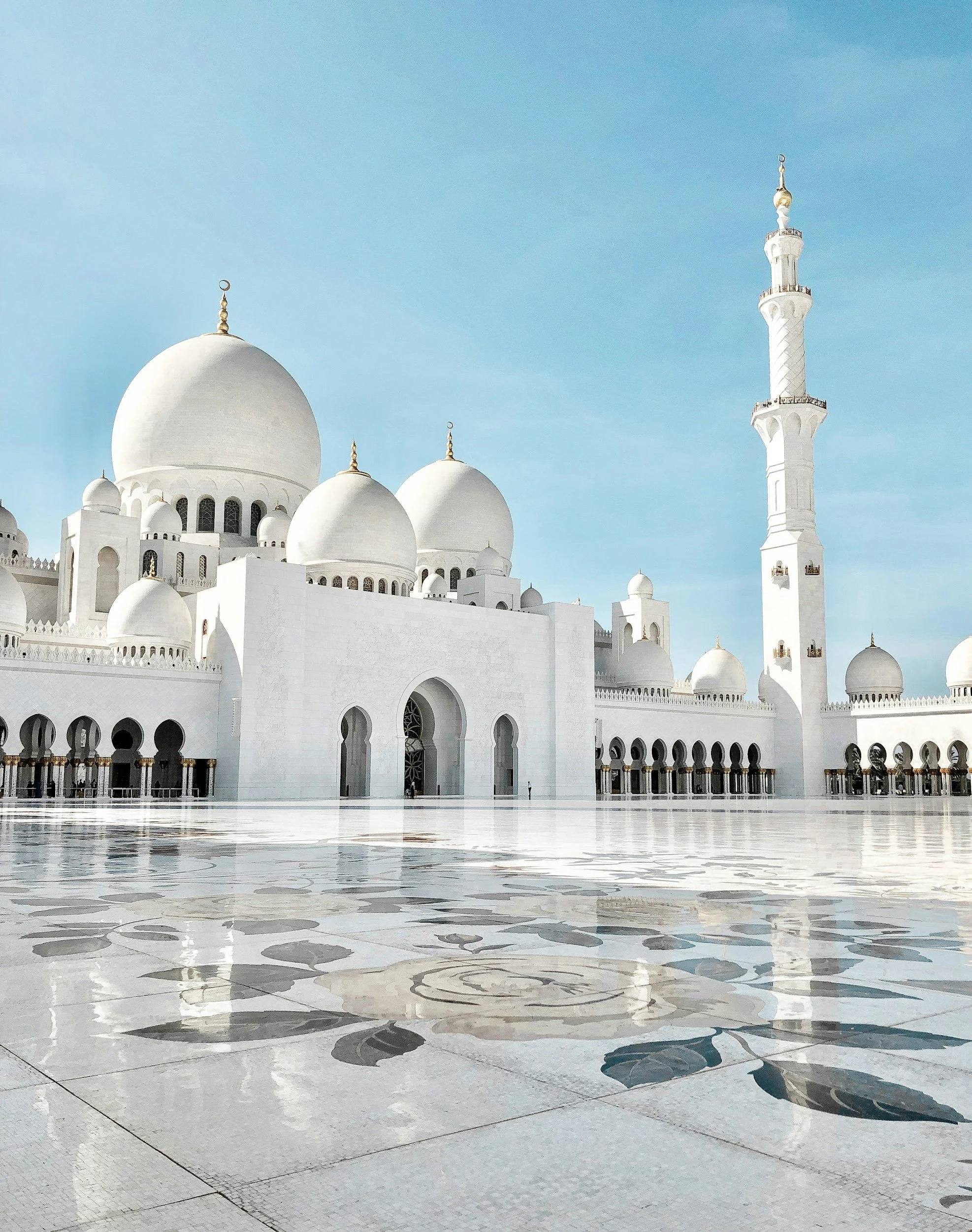 White mosque with multiple domes and a tall minaret against a blue sky, with a reflective tile courtyard in the foreground.