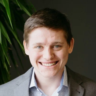 A smiling young man with short brown hair, wearing a grey blazer and a light-patterned dress shirt, standing in front of a dark background with green leaves.
