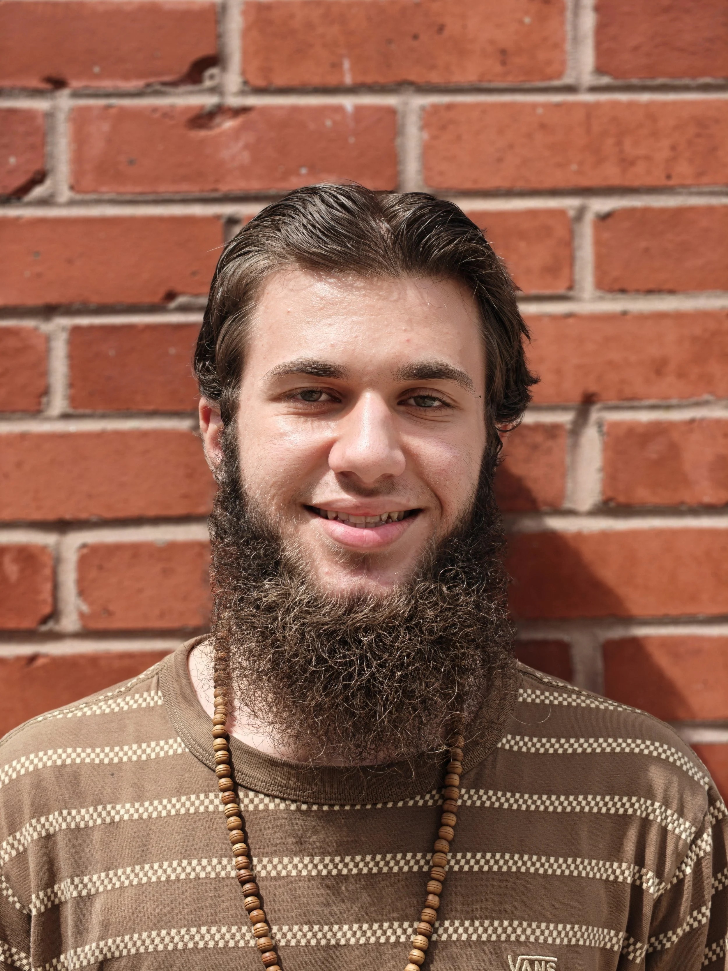 Close-up photo of a young man with a big beard smiling, wearing a beaded necklace and a brown striped shirt, standing against a red brick wall.