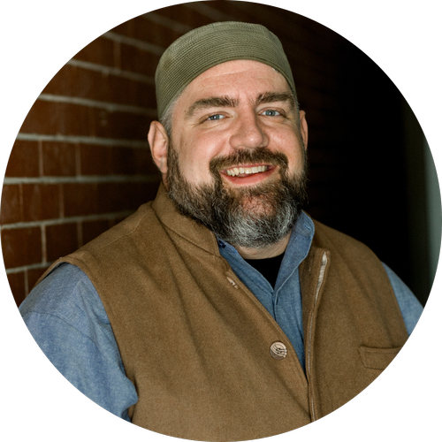 Smiling man with a beard wearing a green beanie and a brown vest, standing against a brick wall.
