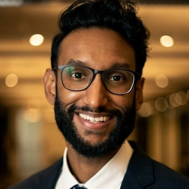 A smiling man with glasses, a beard, and dark hair, wearing a suit and tie, in a warmly lit indoor setting.