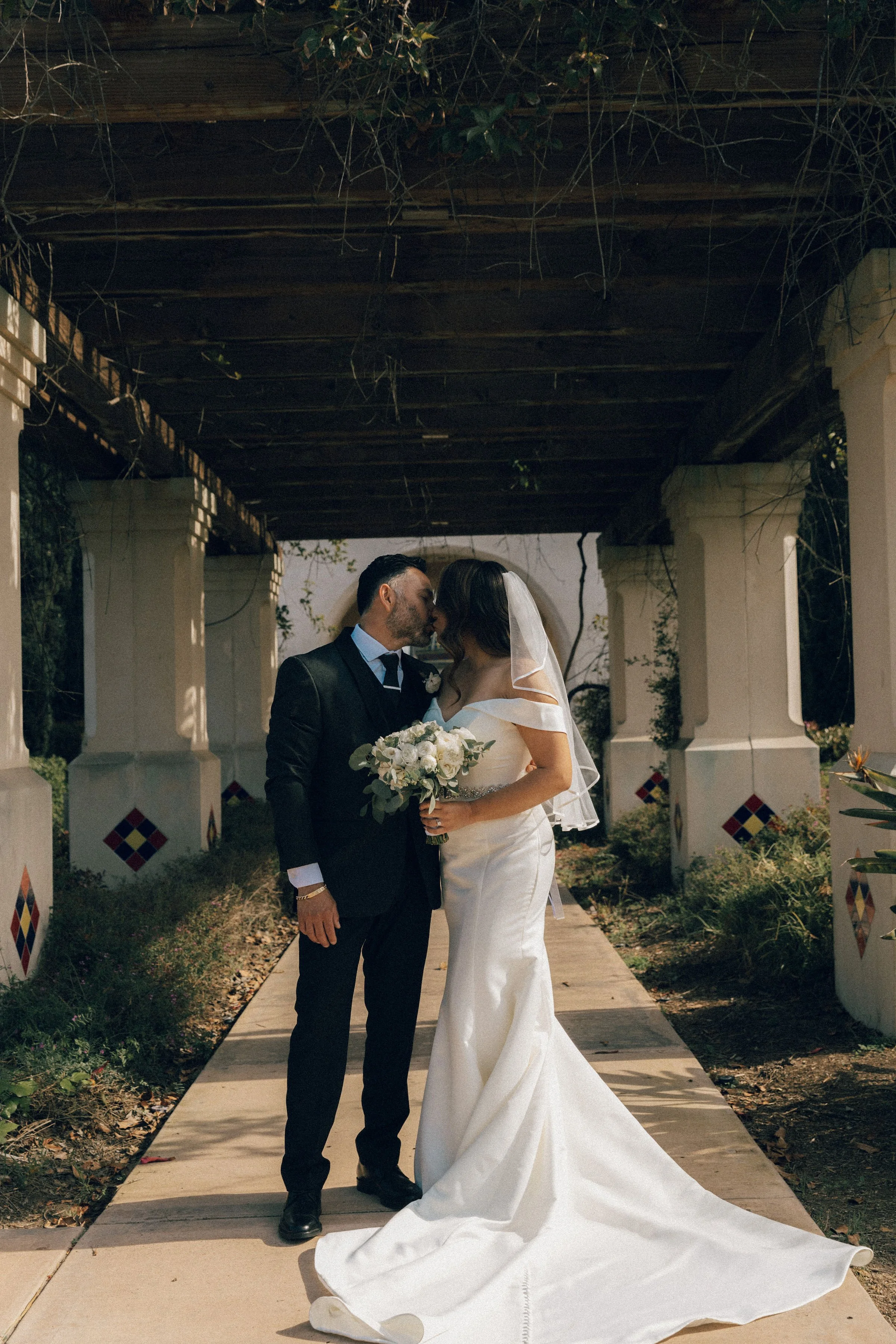 A bride and groom sharing a kiss on their wedding day under a wooden pergola with white columns and colorful tile accents. The bride is holding a bouquet of white flowers, and the couple is dressed in formal wedding attire, with the groom in a black 