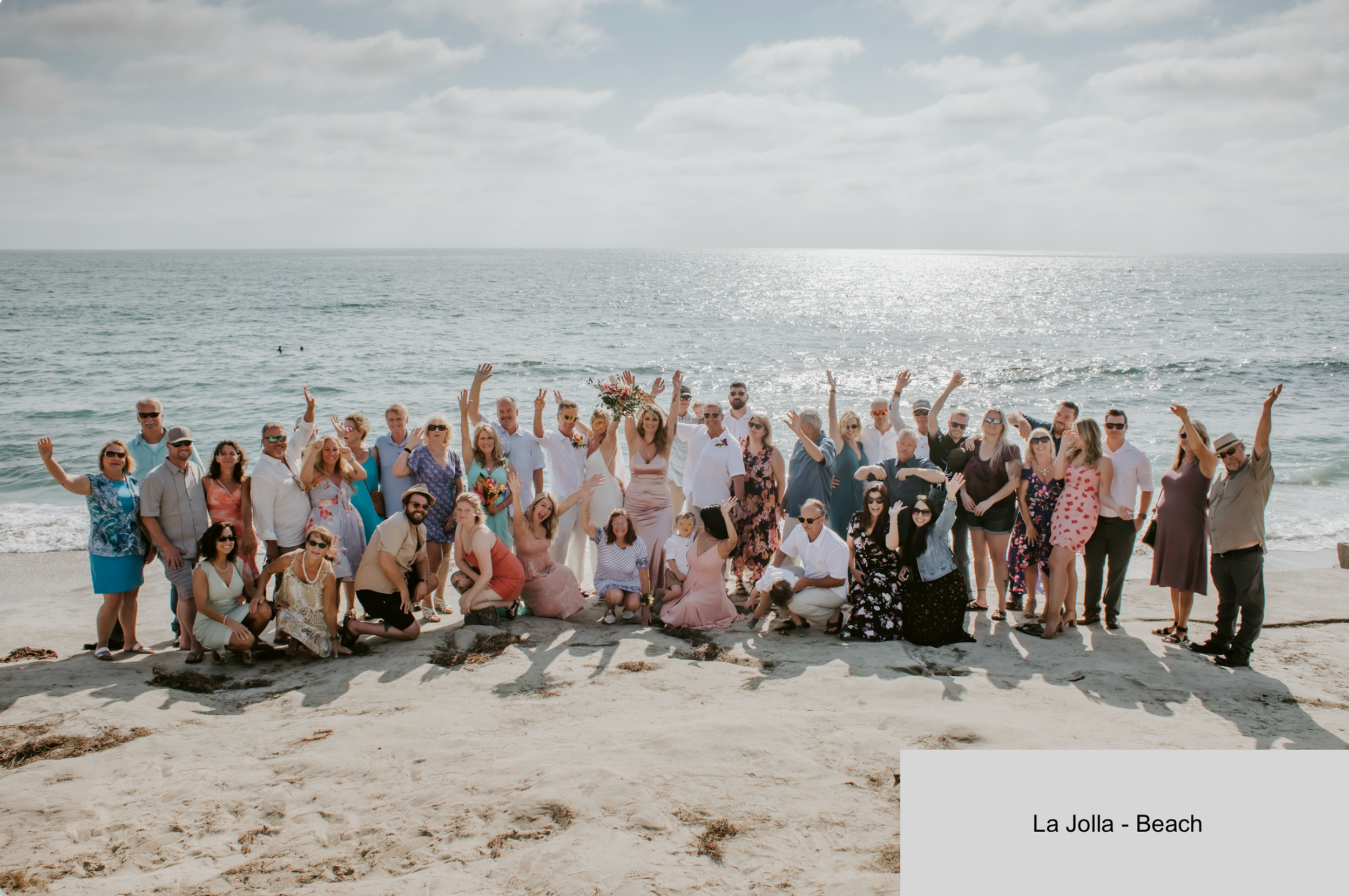 Large group of people gathered on a beach, some are standing and others are kneeling, celebrating. The ocean and cloudy sky are in the background. The scene suggests a beach wedding or celebration at La Jolla Beach.