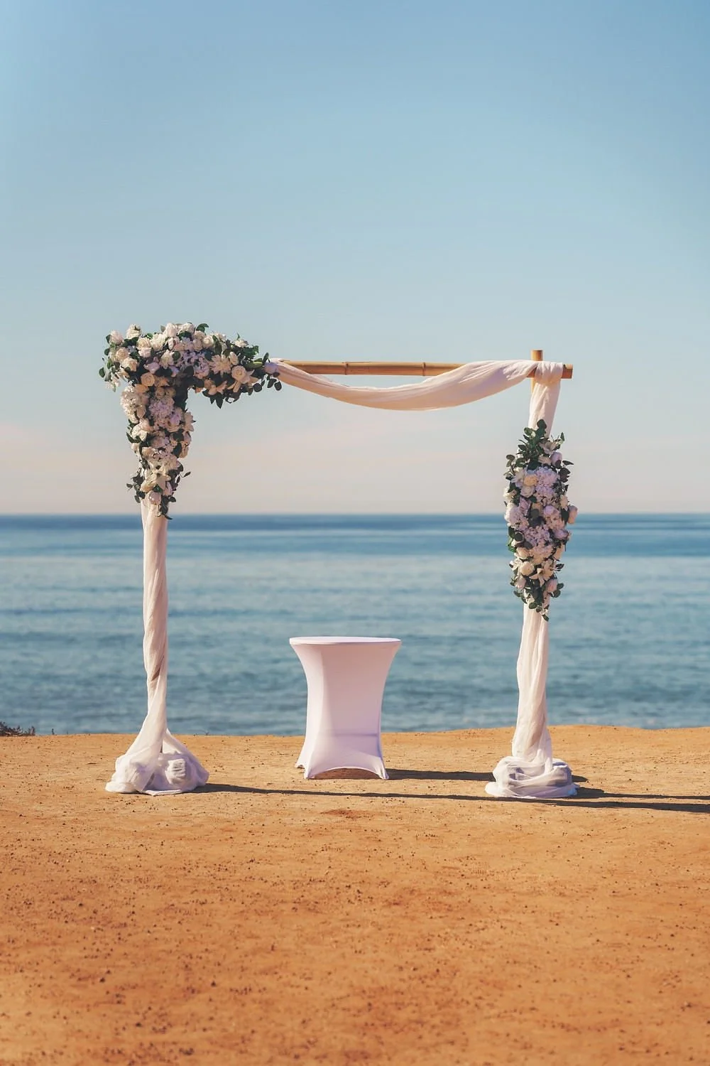 A wedding arch decorated with flowers and draped fabric on a sandy beach with ocean in the background.