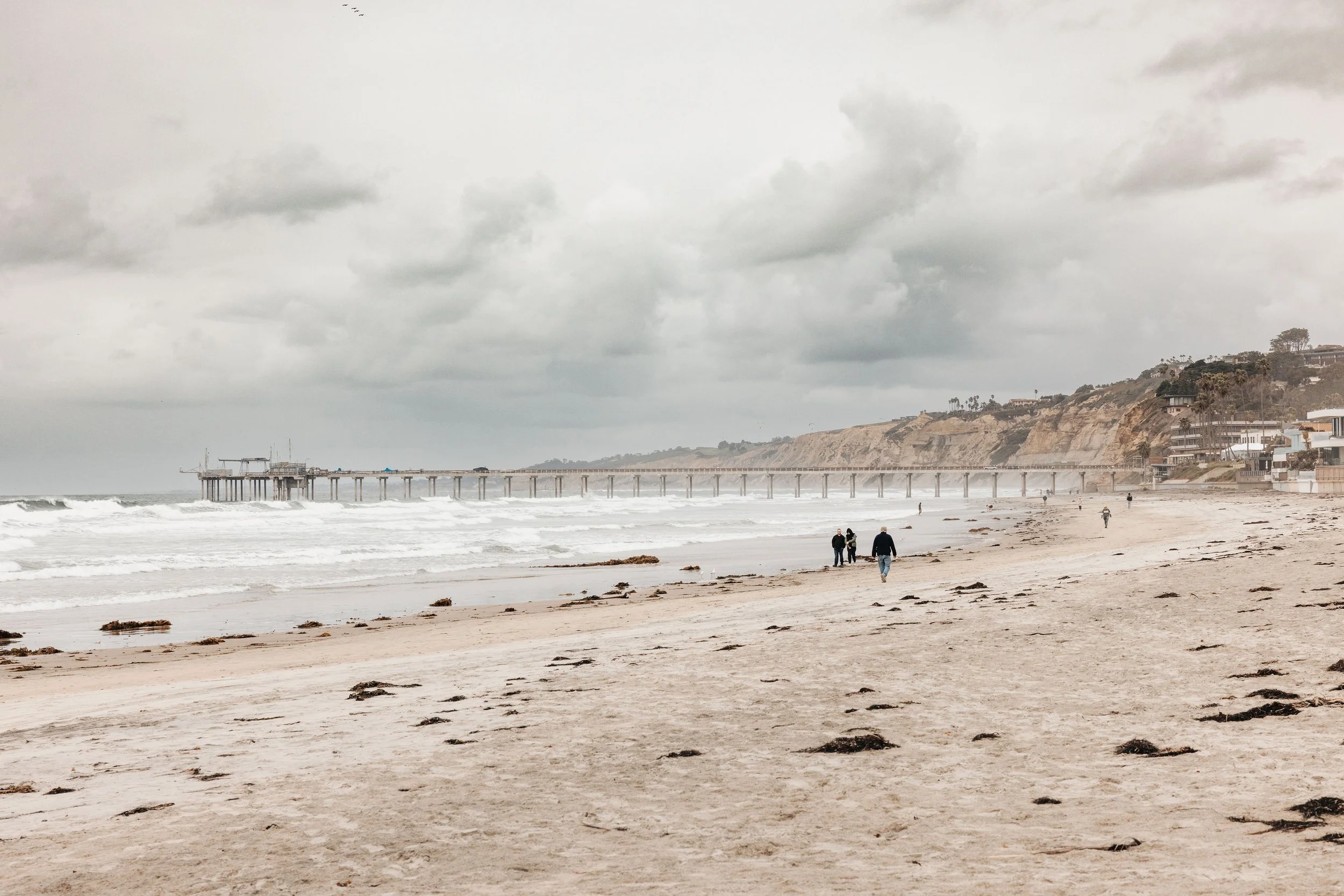 Overcast day at the beach with a pier extending into the ocean, and cliffs with houses in the background. A few people walk along the sandy shore.