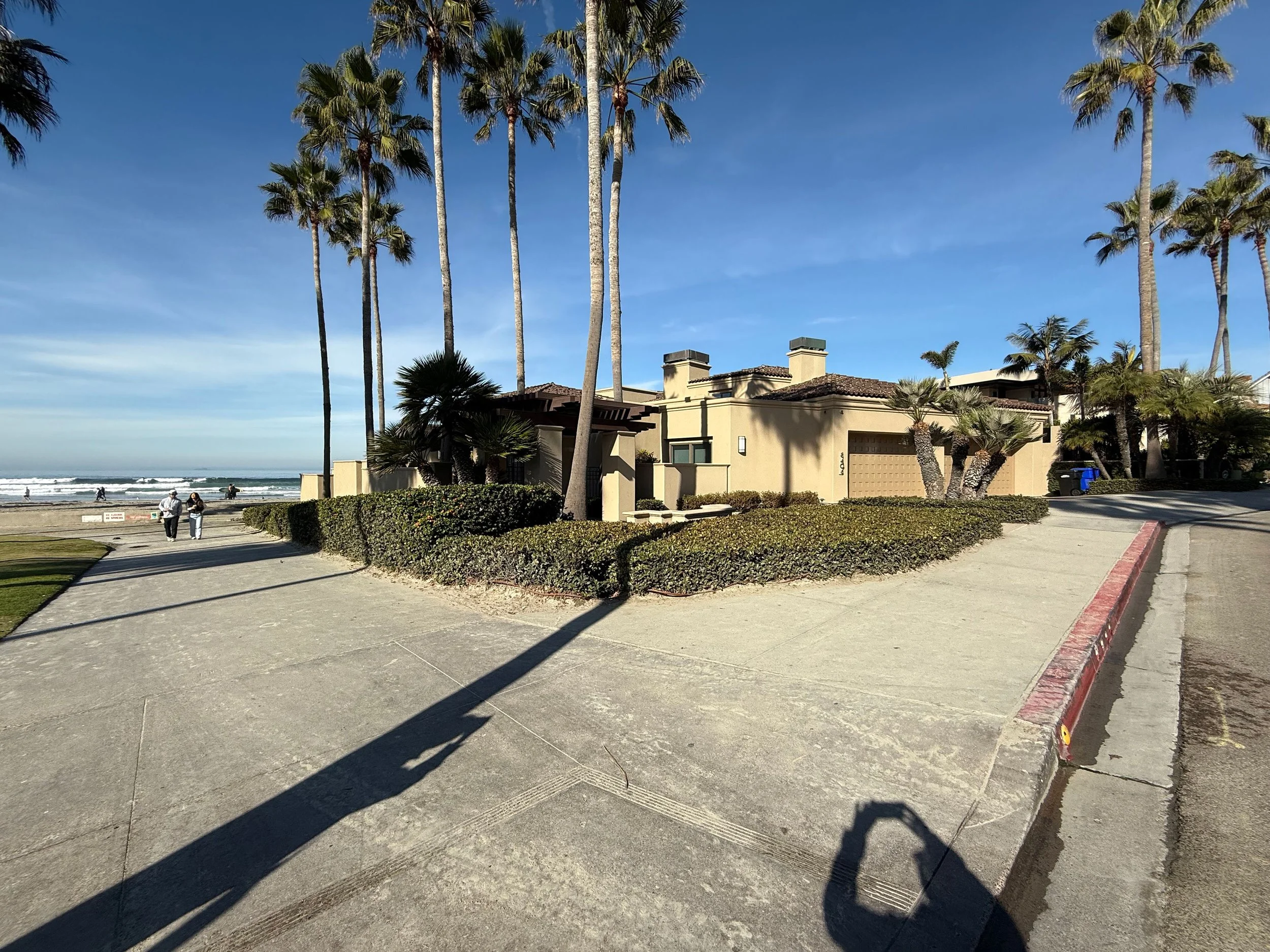 Beachfront house with tall palm trees and a sandy pathway leading to the beach, visible waves and people in the distance, clear blue sky.