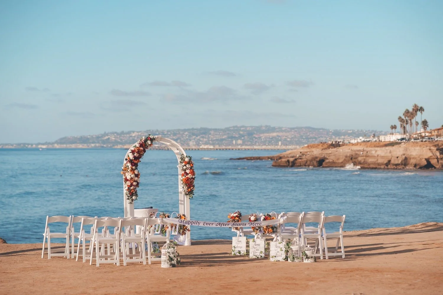 Beachside wedding altar decorated with flowers and white chairs, overlooking ocean and coastline with palm trees.