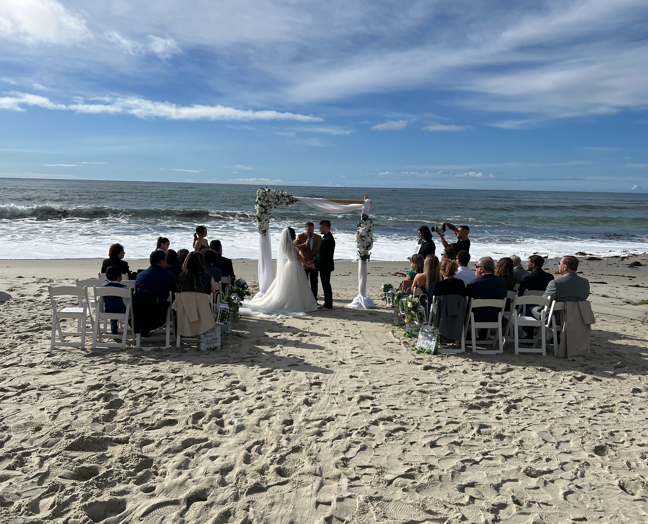 A beach wedding ceremony setup with white chairs, floral decorations, and an arch with white drapery and flowers, facing the ocean under a partly cloudy sky.