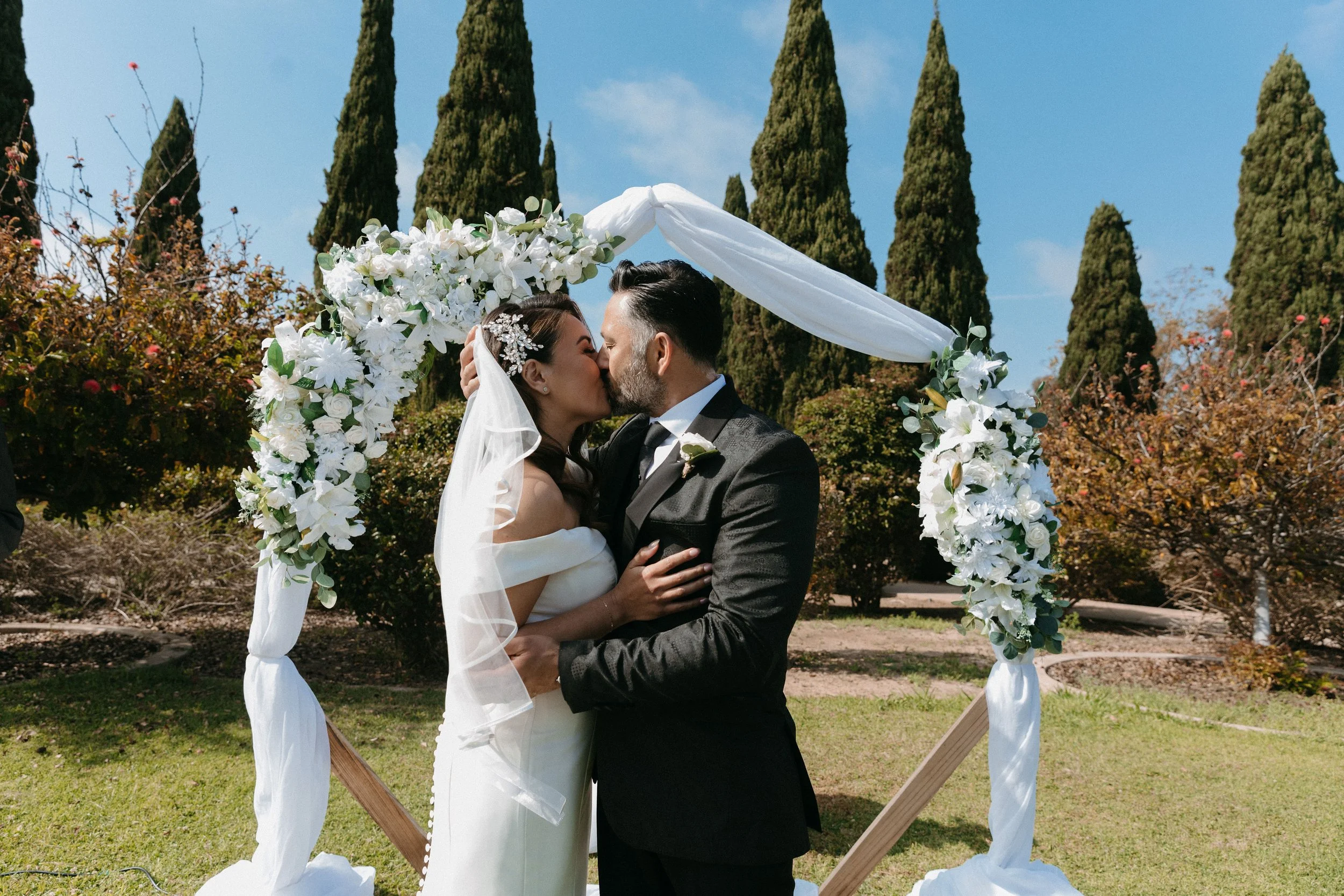 A bride and groom kissing under a floral wedding arch outdoors, with tall cypress trees in the background.