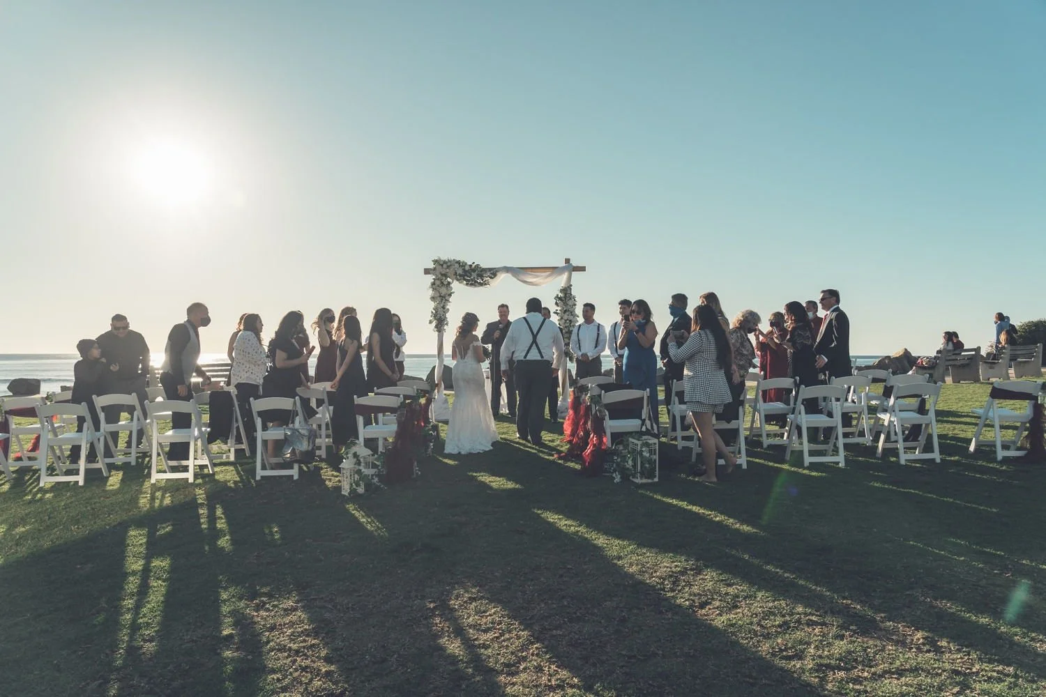 A group of people celebrating a wedding outdoors near the beach, with the bride and groom standing under a decorated arch. Chairs are arranged in rows on the grass, and wedding guests are dressed in formal attire, some wearing masks. The sun is low i