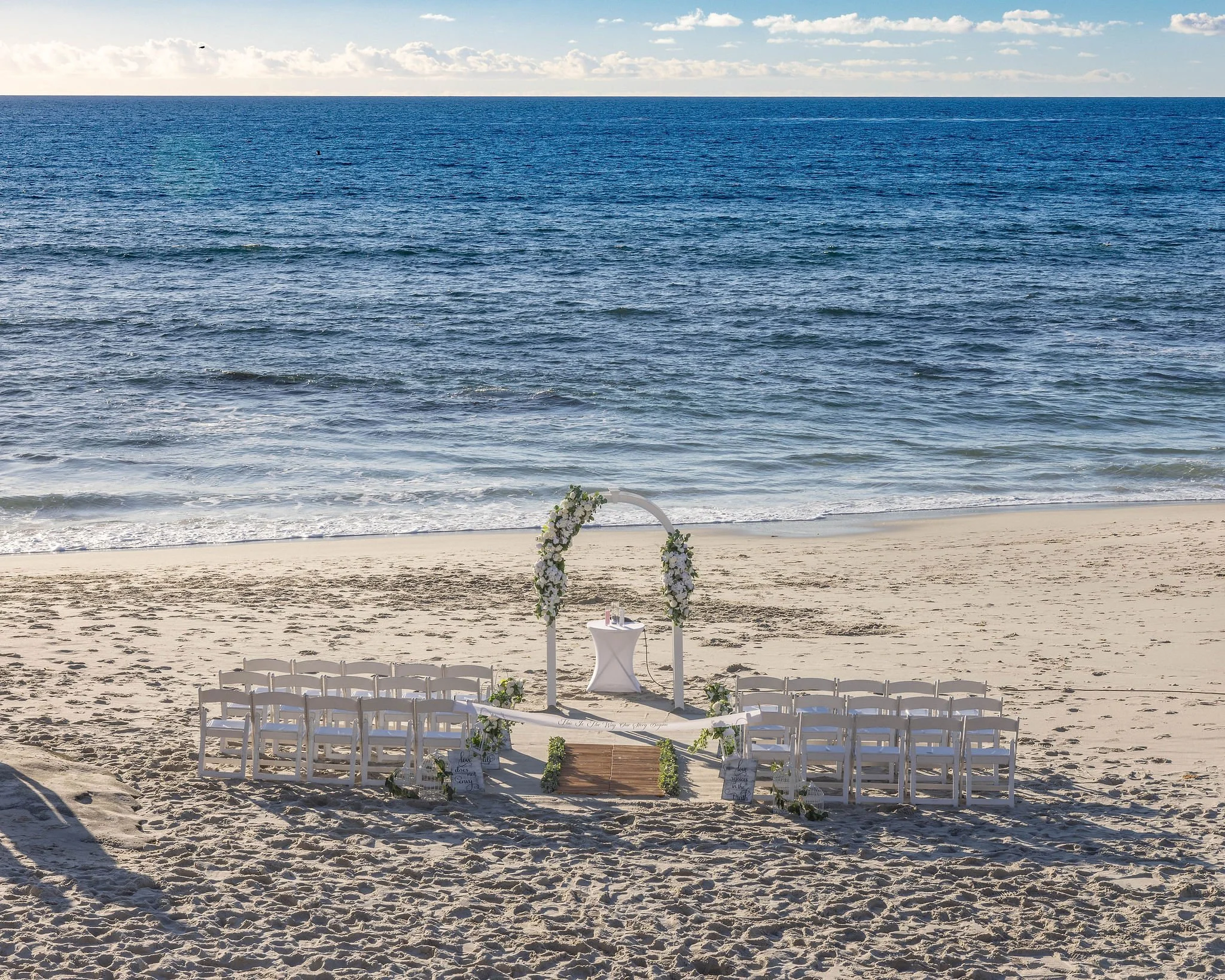 A wedding ceremony setup on a sandy beach with chairs arranged facing an arch decorated with white flowers, overlooking the ocean under a partly cloudy sky.