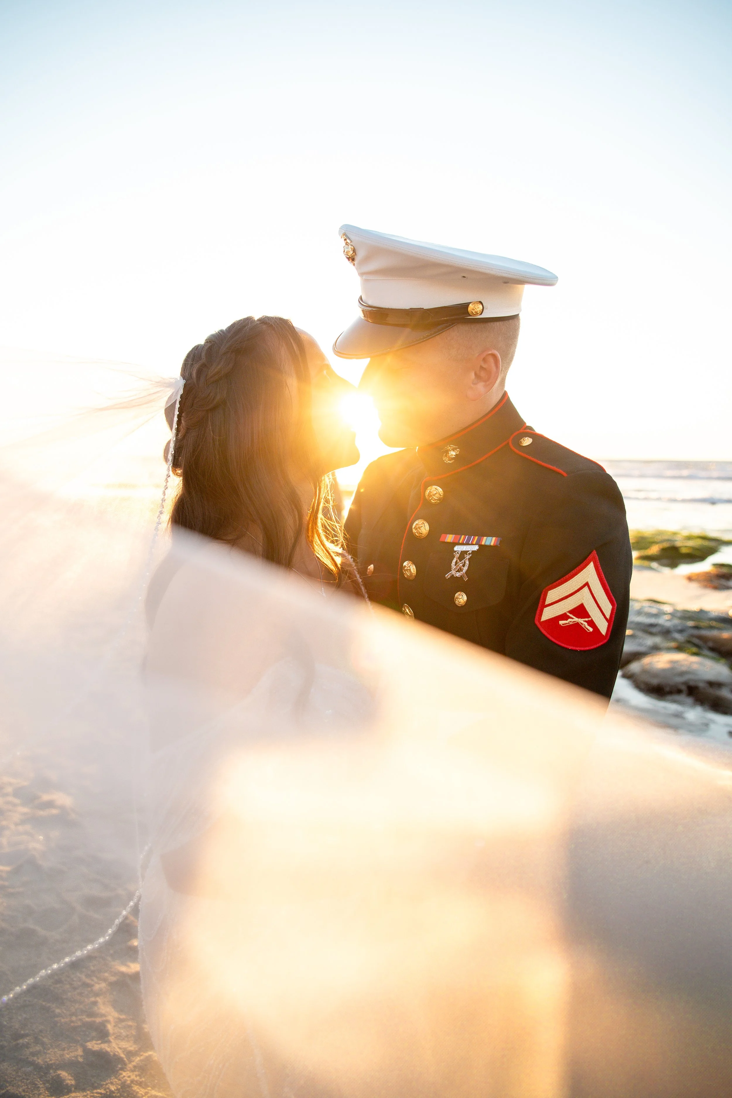 A couple is kissing on a beach at sunset. The man is wearing a U.S. Marine Corps dress uniform, and the woman has long brown hair and is wearing a white dress with a veil. The sun is setting behind them, casting a warm glow.