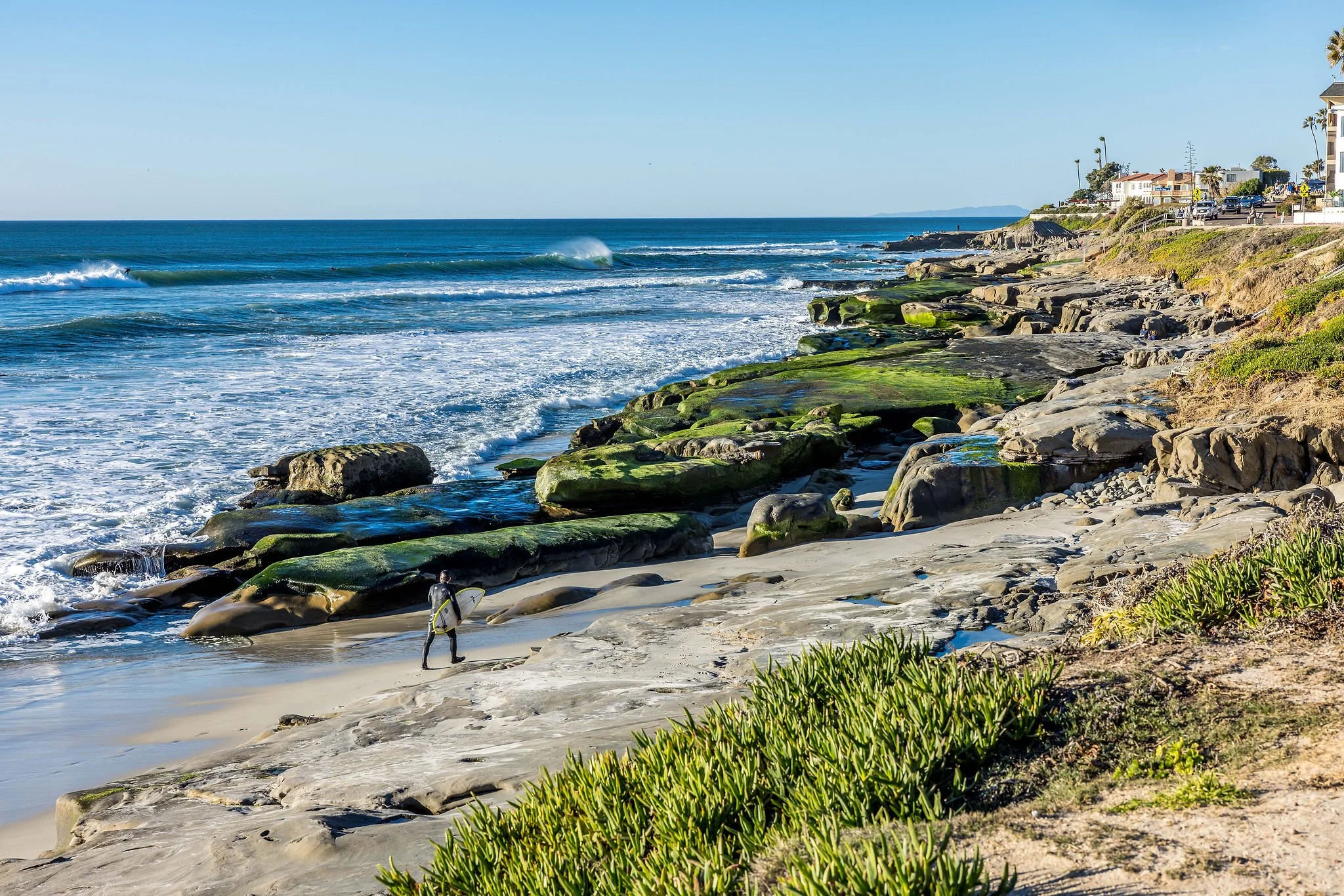 A person with a surfboard walking on the sandy beach near the shoreline, with waves in the ocean and houses on the hill in the background.