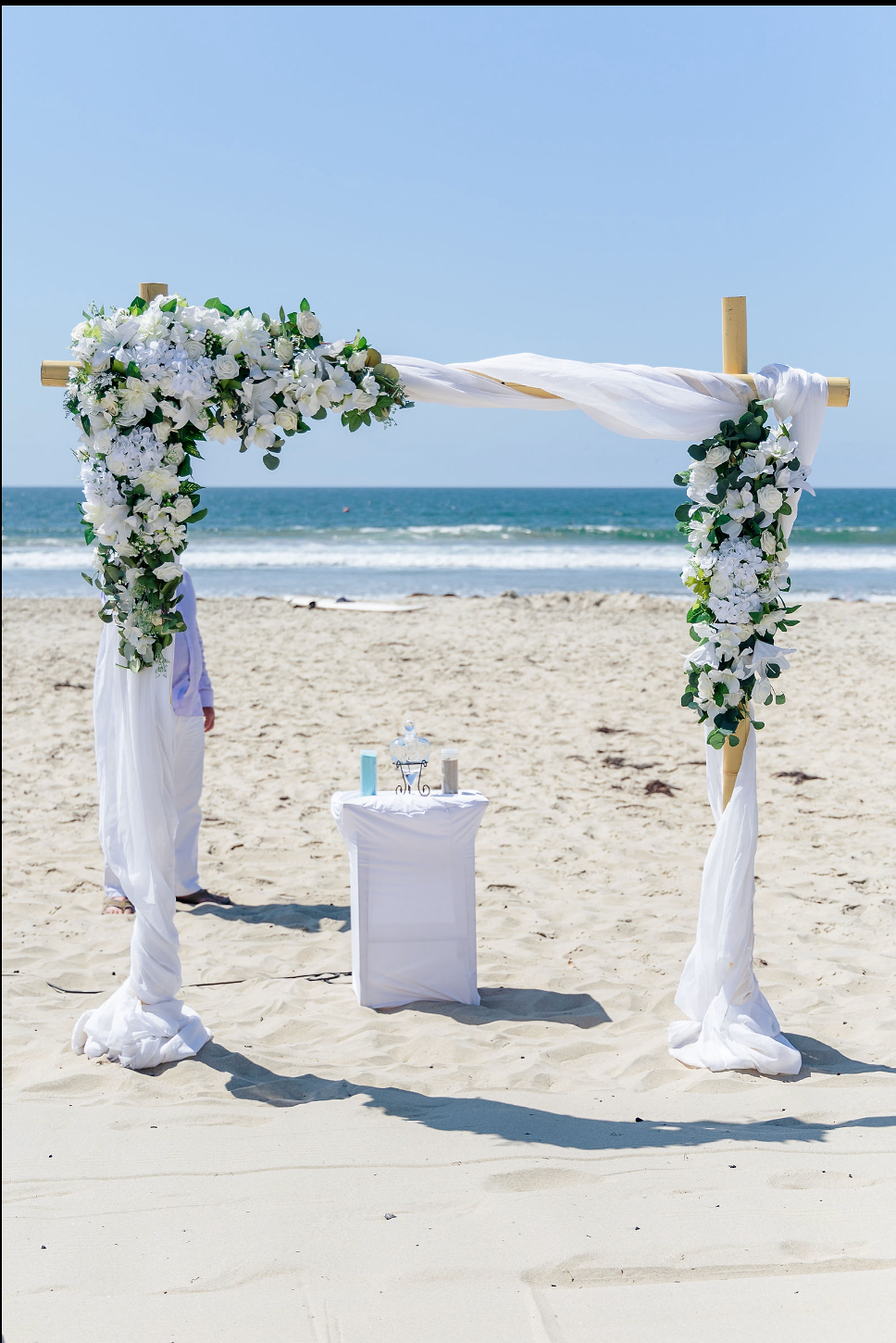 A wedding arch decorated with white flowers and green leaves set up on a sandy beach with the ocean in the background.
