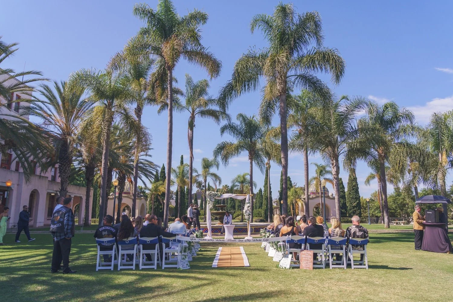 An outdoor wedding ceremony with white chairs arranged on a grassy lawn, palm trees, and a fountain in the background under a clear blue sky.