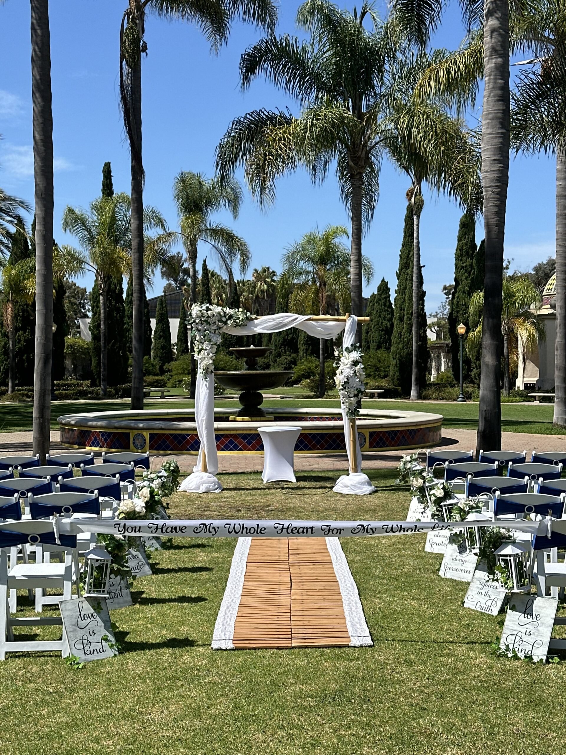 Outdoor wedding ceremony setup with a wooden aisle runner, white chairs, floral decorations, and an arch with draped white fabric and flowers, surrounded by tall palm trees and greenery on a sunny day.