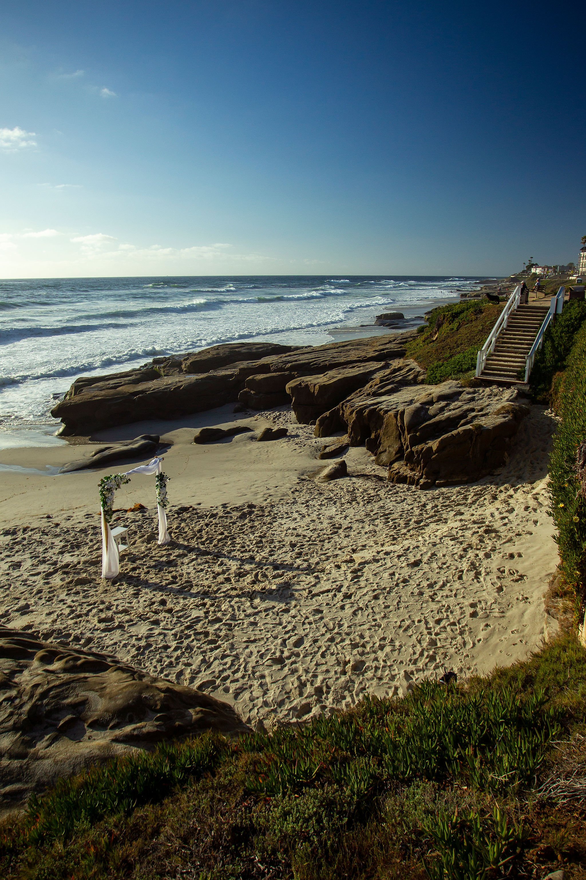 Beach with rocks, ocean waves, a clear sky, and stairs leading to the walkway.