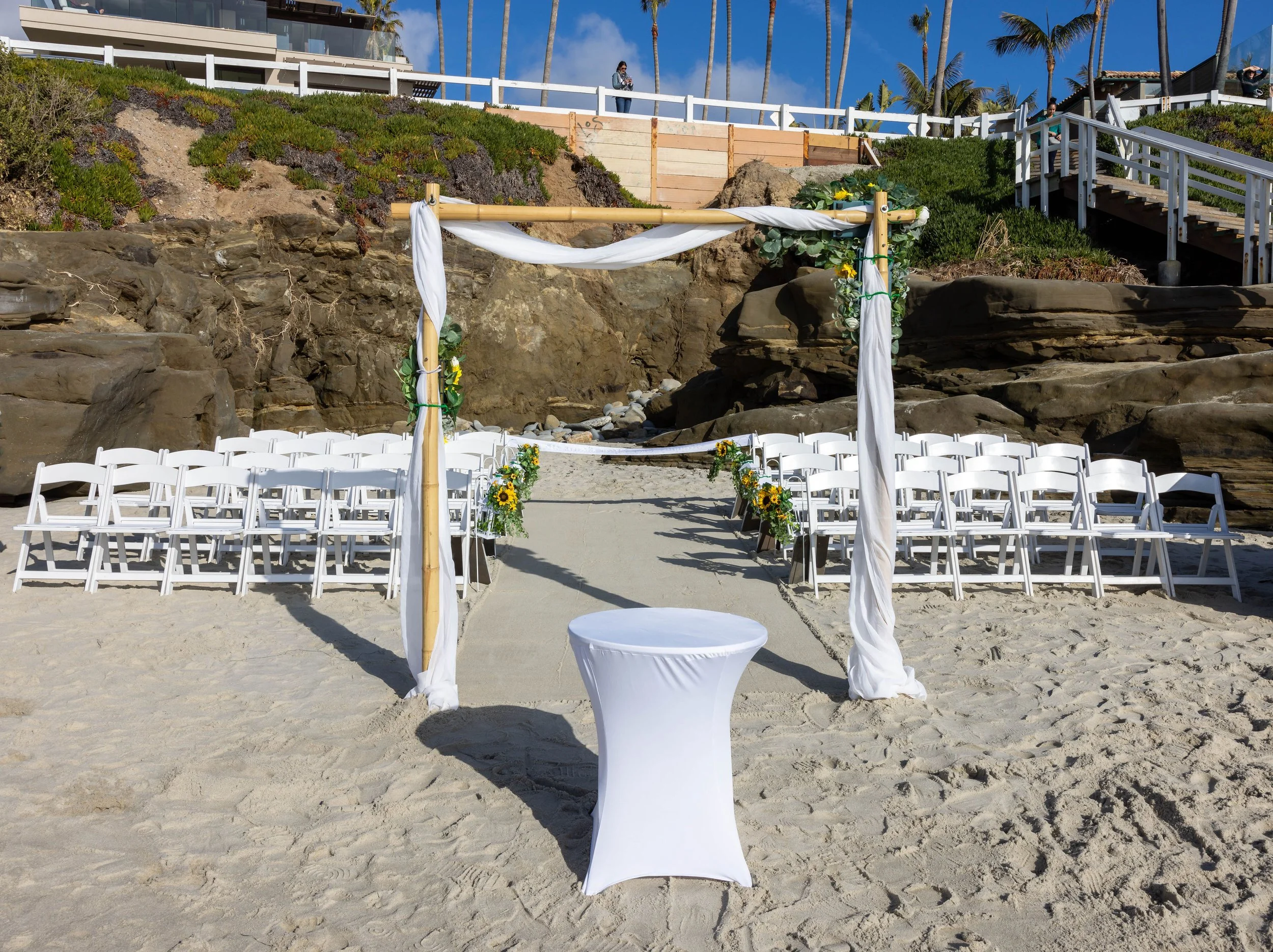 Beachside wedding setup with a white draped arch decorated with flowers, overlooking rocky cliffs. Rows of white chairs are arranged for guests, with a small round table in front of the arch. Stairs and people are visible in the background near the c