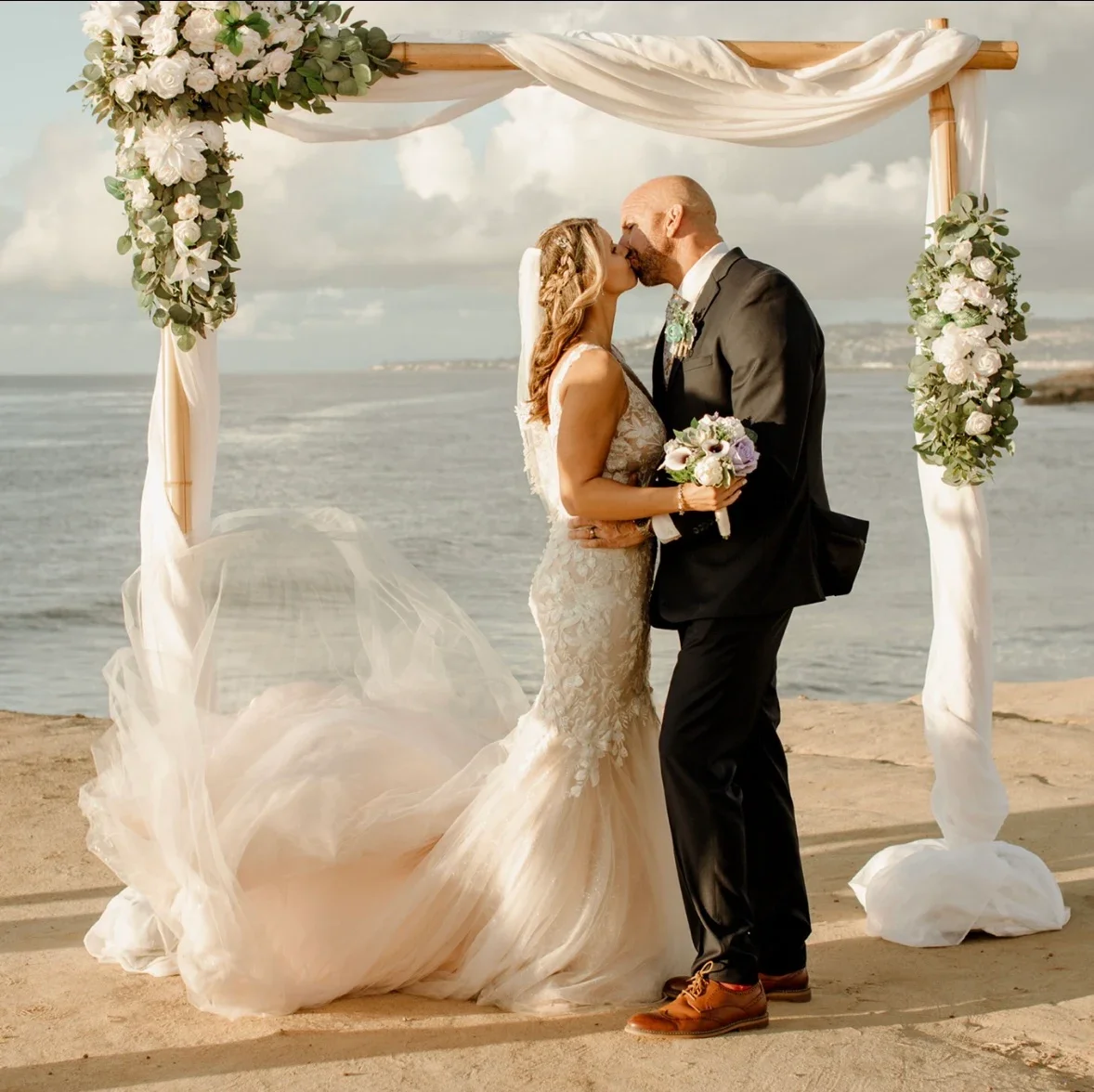 A bride and groom kissing during their beach wedding ceremony beneath a floral arch with white roses and greenery, ocean in the background.